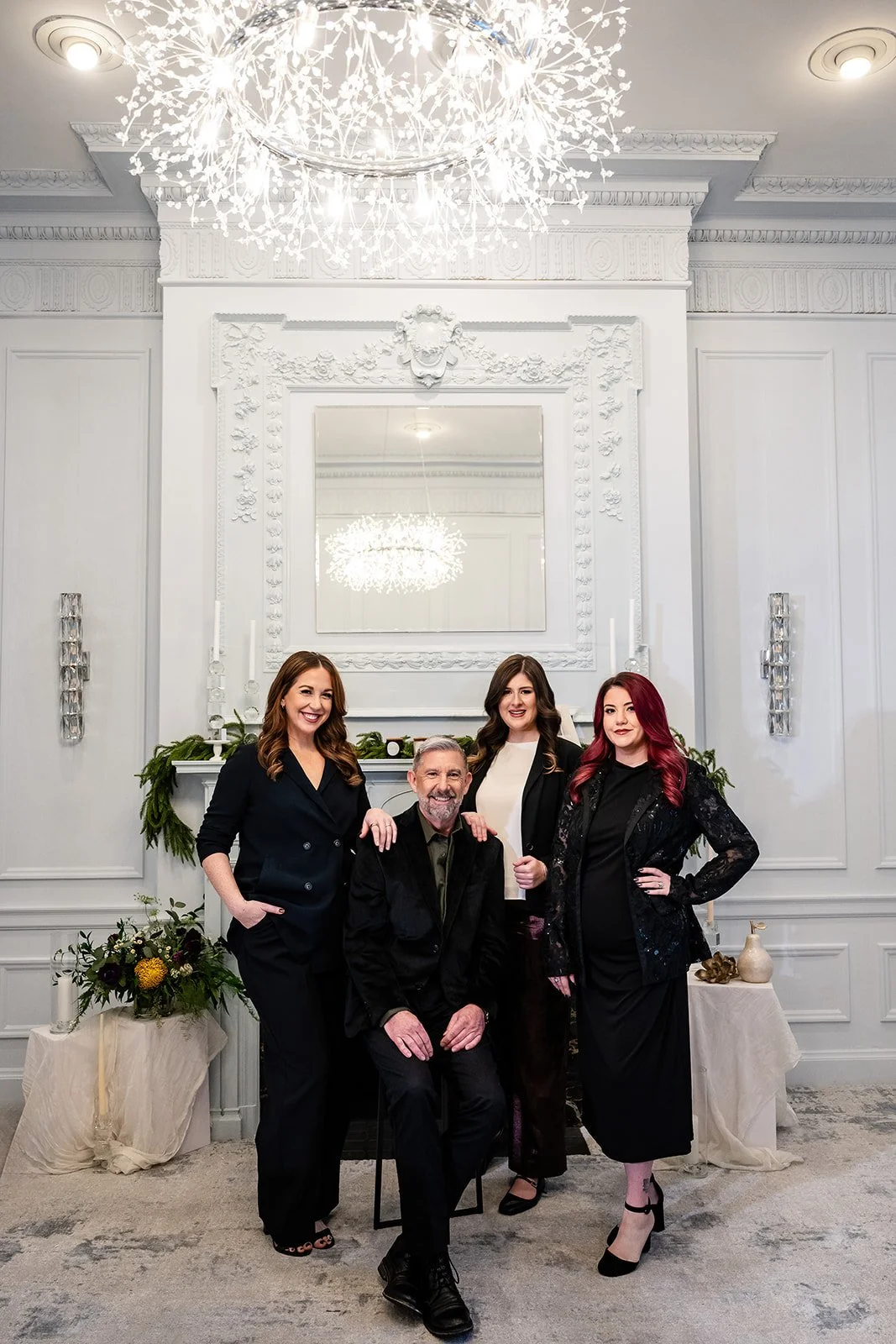 Three women in black standing around a man in black sitting in front of a fireplace