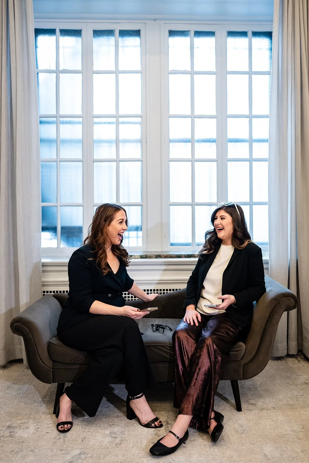 two women sitting on couch holding phones and laughing