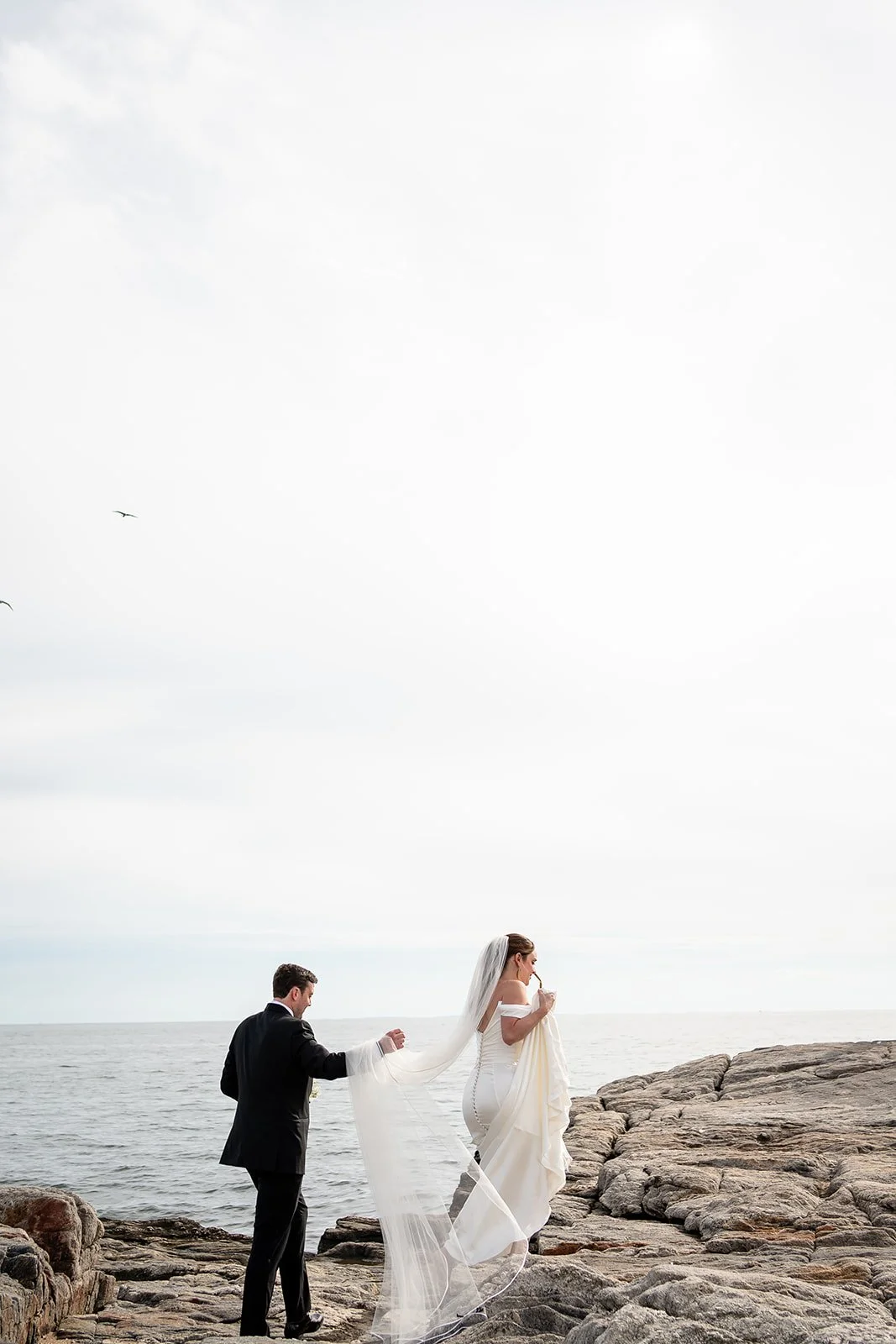 woman in wedding dress walking up rocks with a man following behind carrying her veil