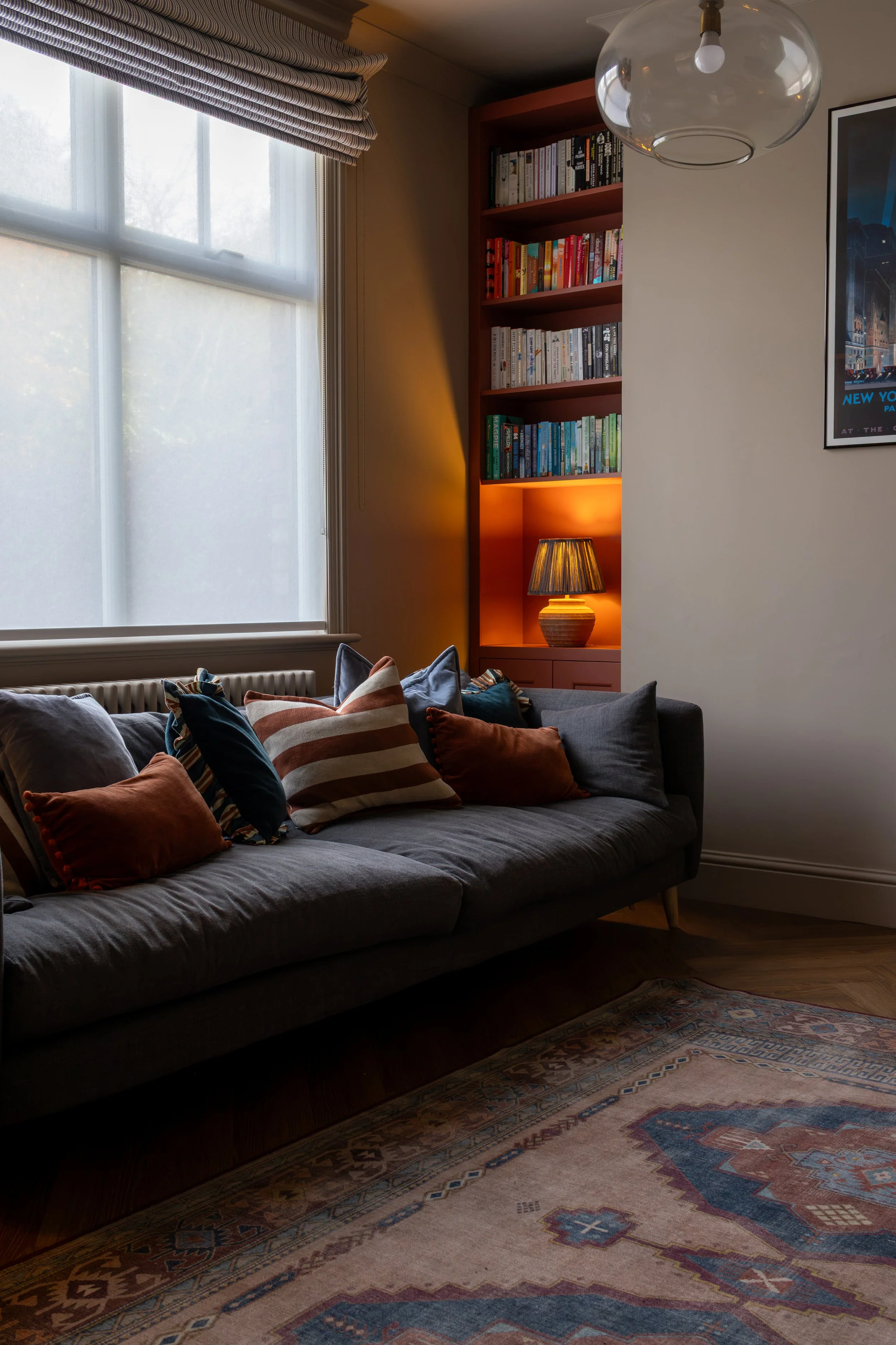 Grey sofa piled with terracotta and blue cushions beside a glowing alcove bookshelf lamp
