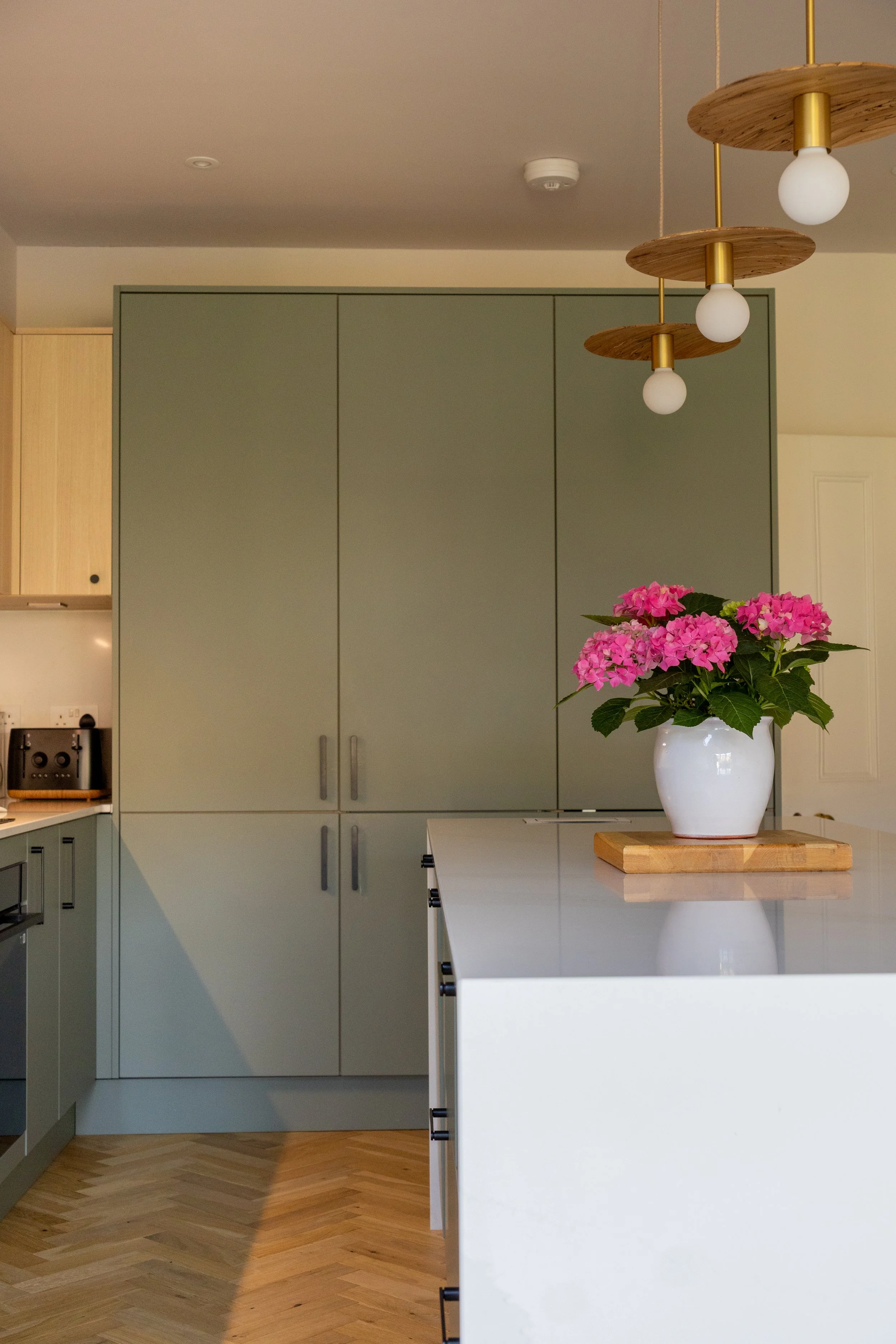 Tall sage green kitchen cabinetry with wood and brass disc pendant lights and pink hydrangeas
