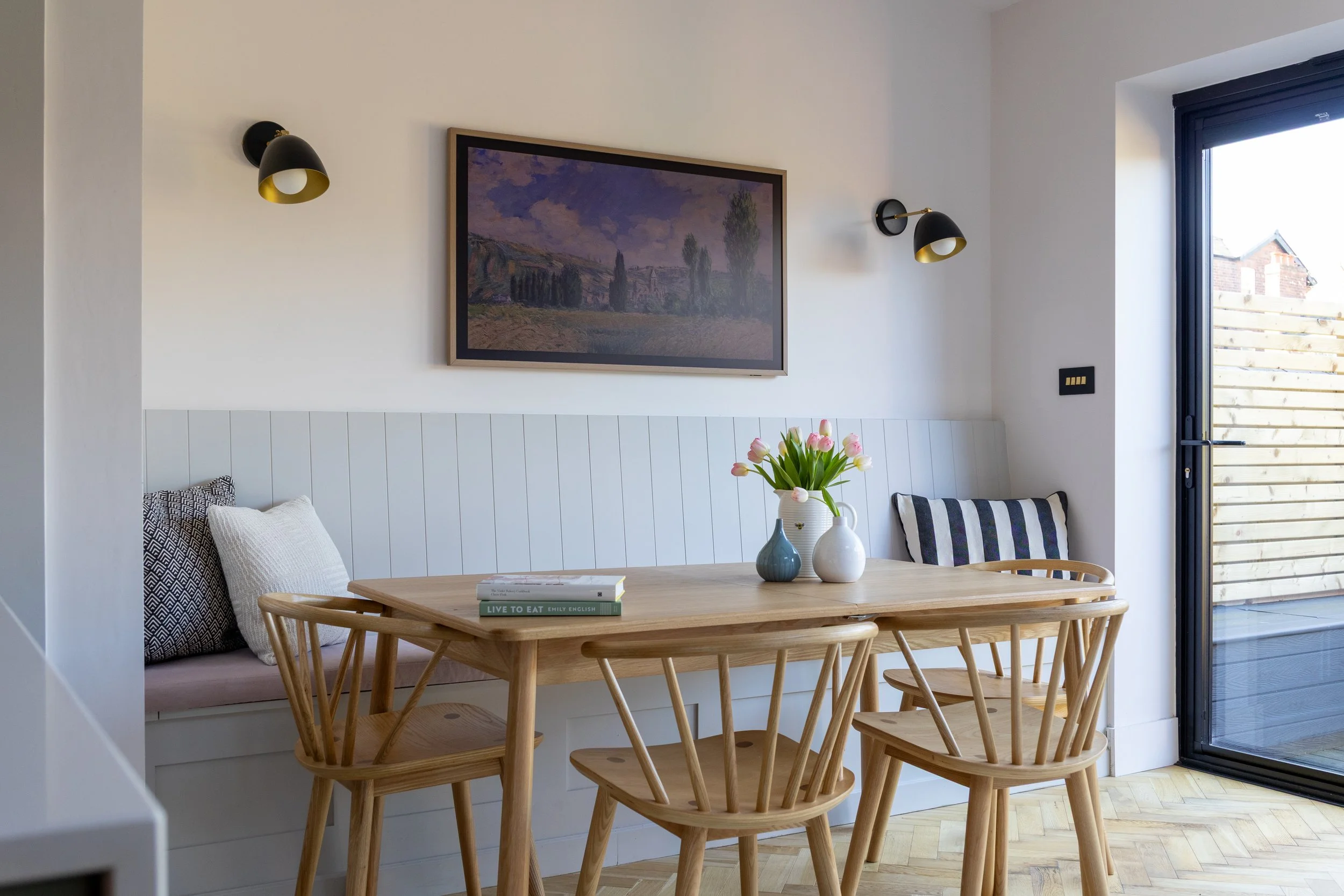 Oak dining table with pink tulips in a jug beside a framed landscape painting and striped cushion