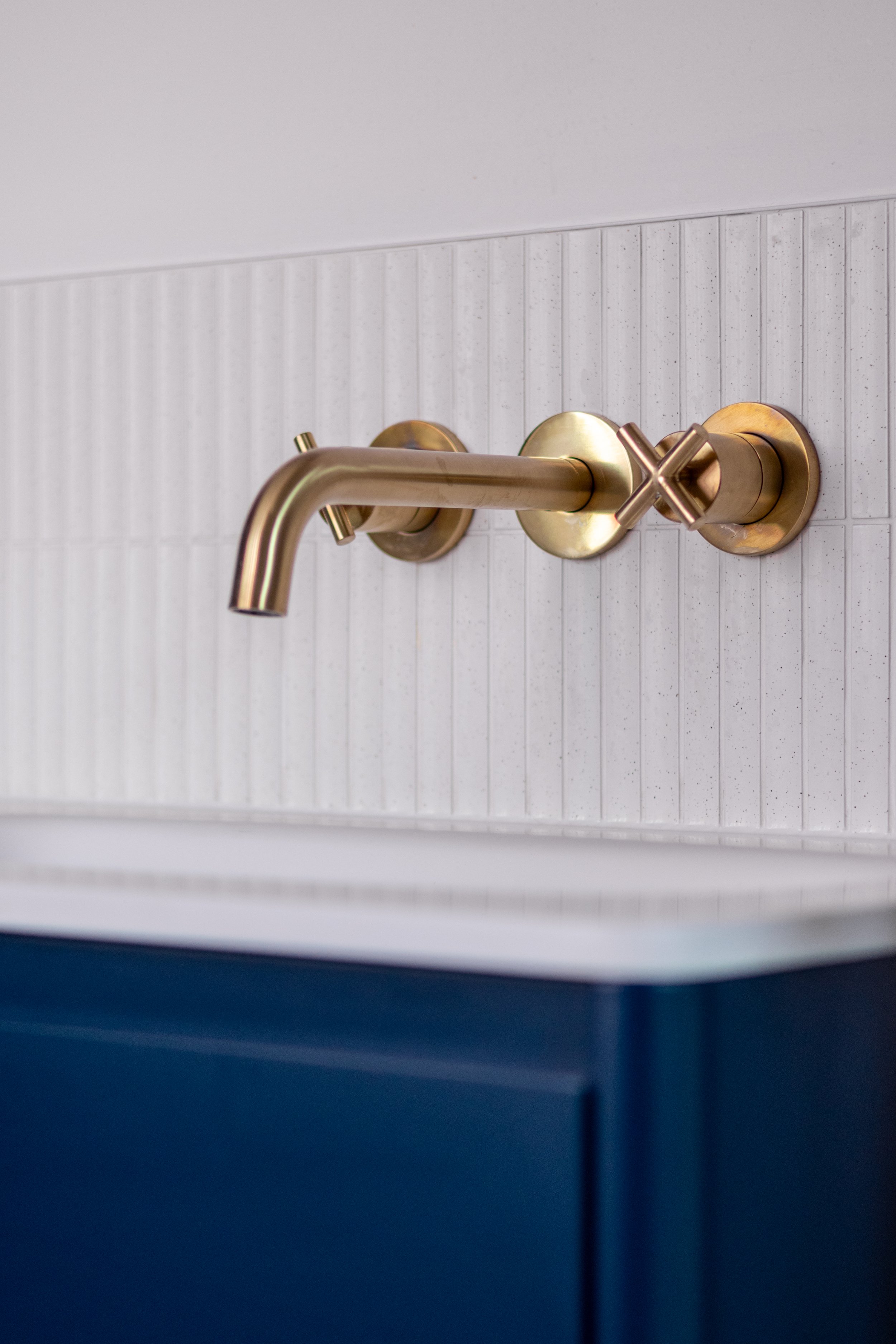 Brass wall-mounted basin tap with cross handle above a navy vanity unit