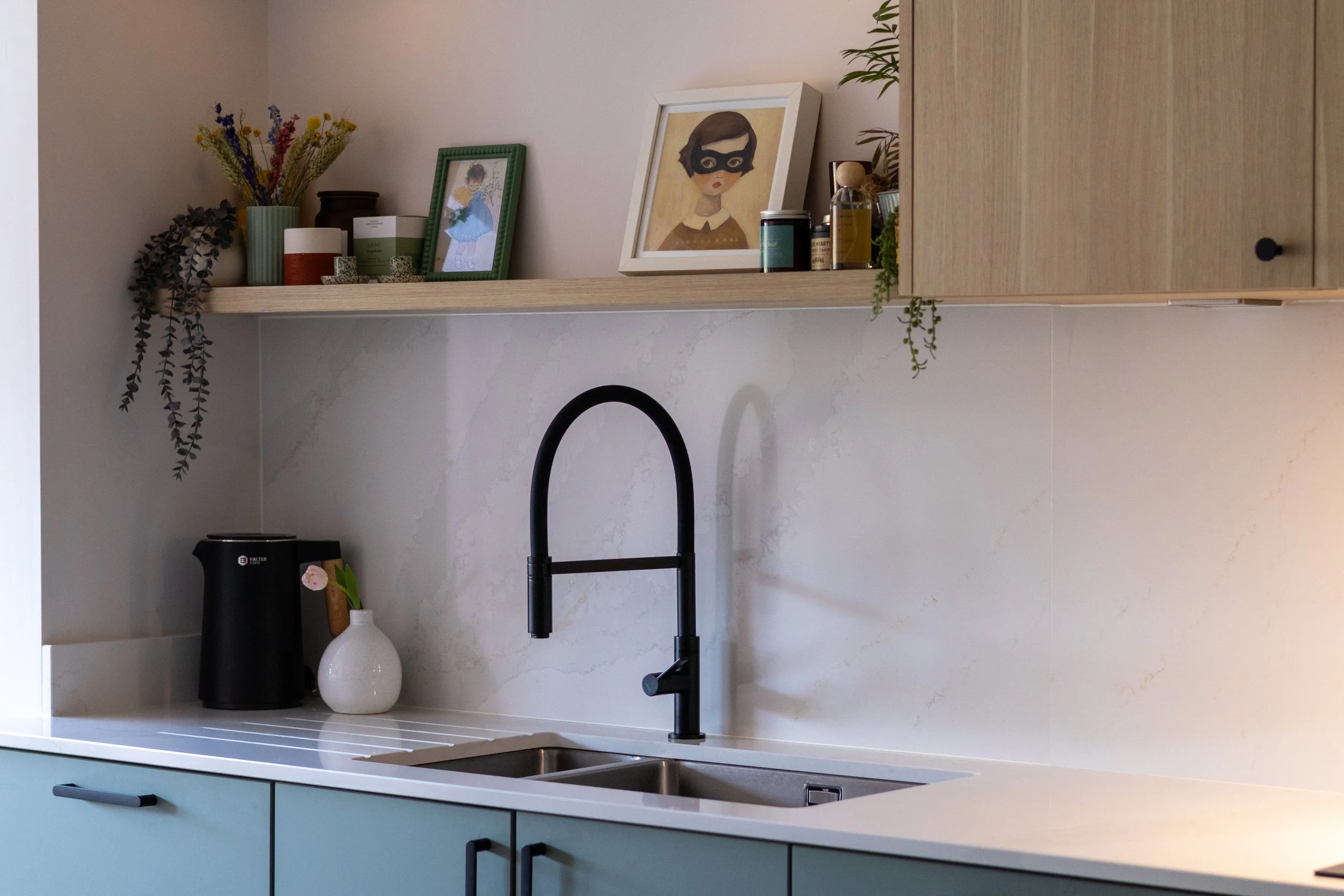 Kitchen sink area with matte black tap, oak floating shelf and framed illustrations