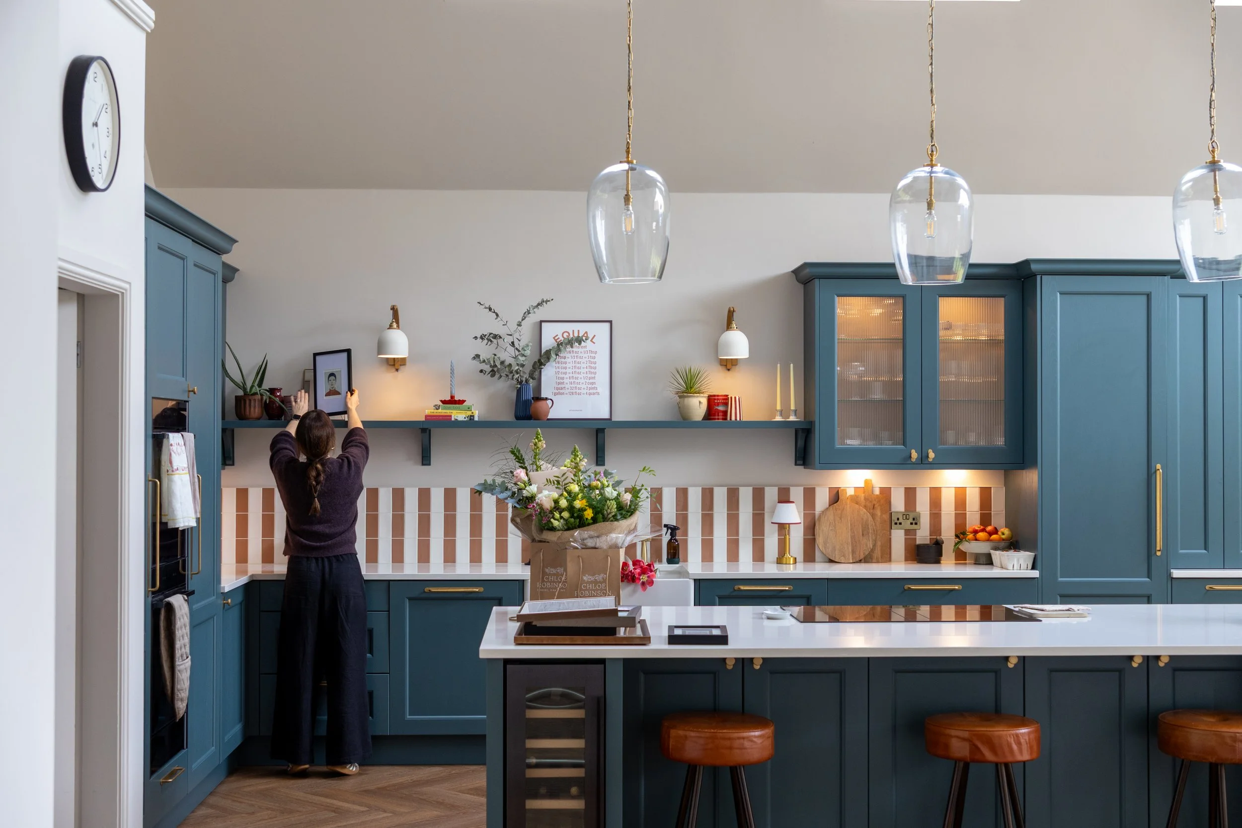 Woman arranging a framed print on an open shelf in a teal shaker kitchen with striped tile splashback