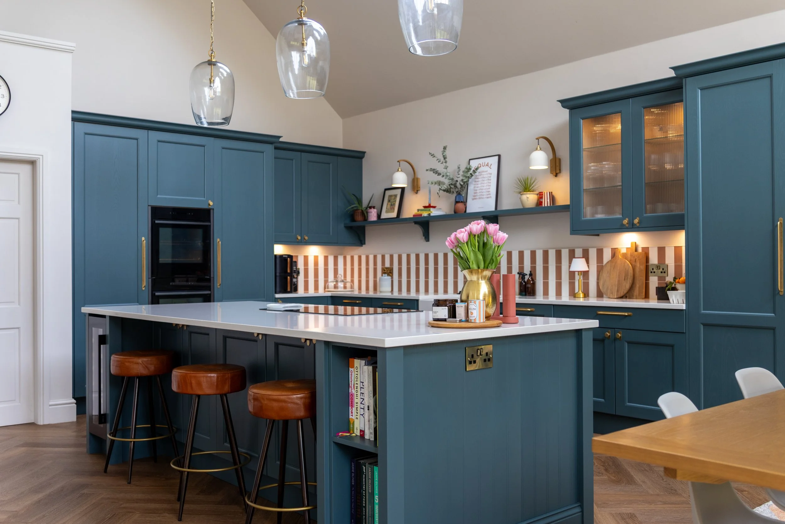Teal kitchen island with tan leather stools, glass pendant lights and built-in bookshelf