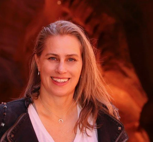 A smiling woman with long blonde hair, wearing a black jacket over a light-colored shirt, standing outdoors with a reddish rock formation in the background.