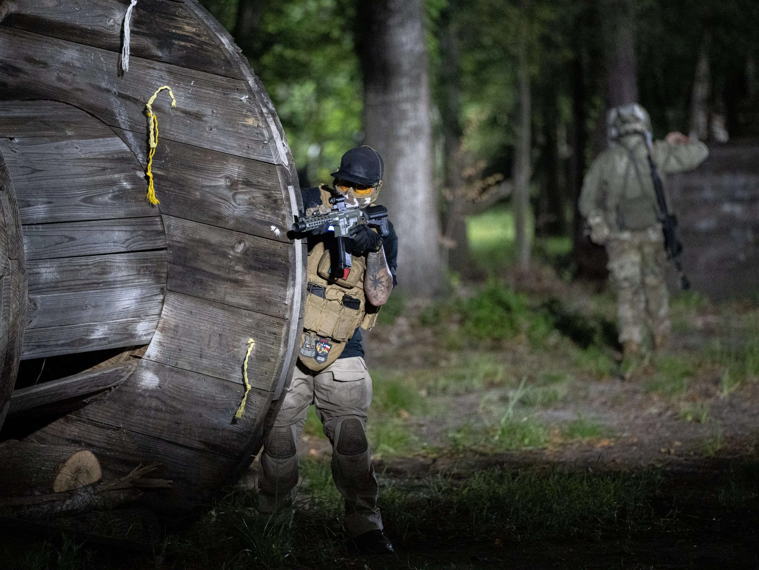 A soldier in tactical gear aiming a rifle around a corner of a wooden structure in a forested area at dusk.
