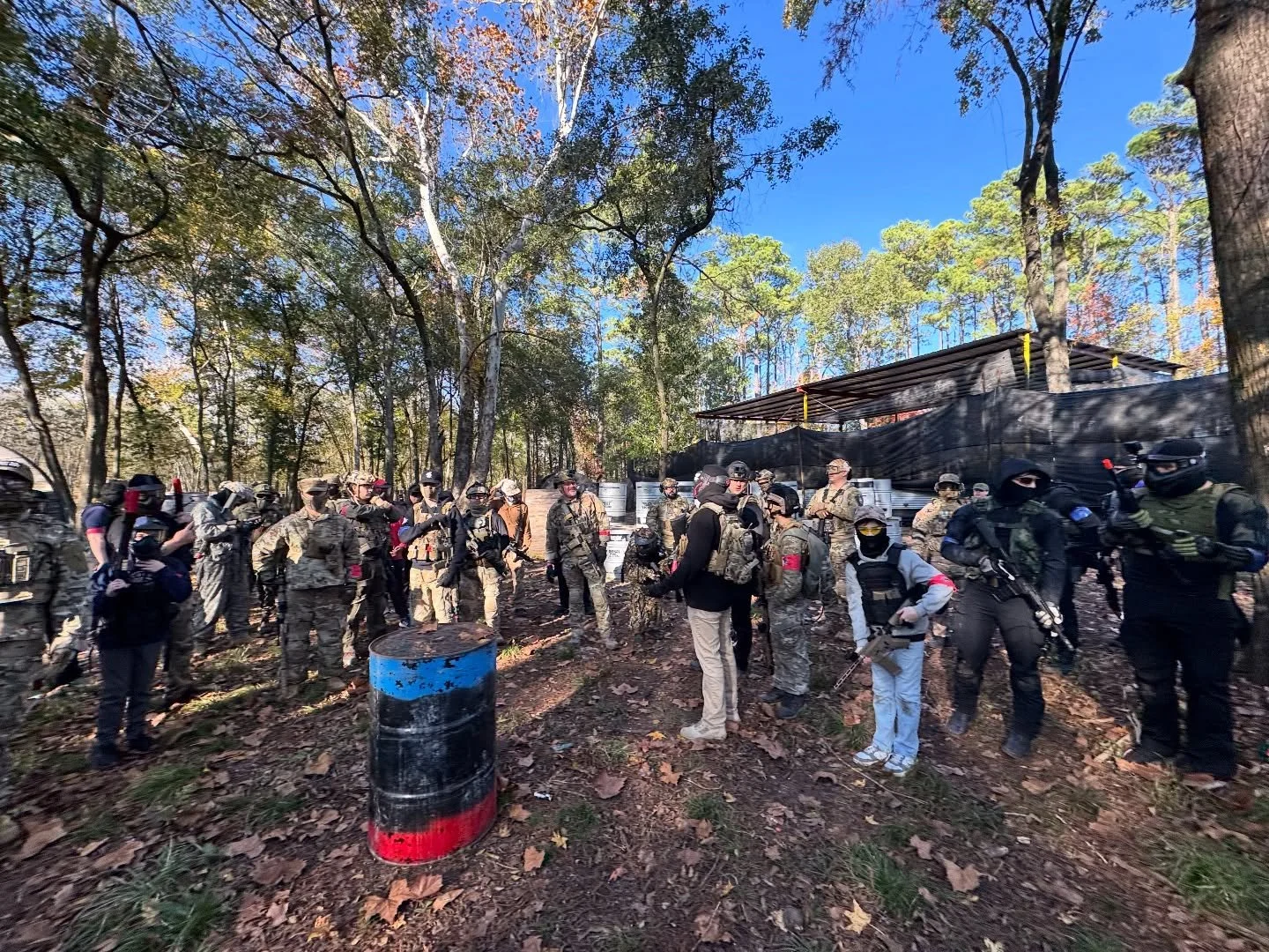 Group of people in tactical gear gathered outdoors in a wooded area during daytime, some holding airsoft guns, with a makeshift shelter in the background and fallen leaves on the ground.