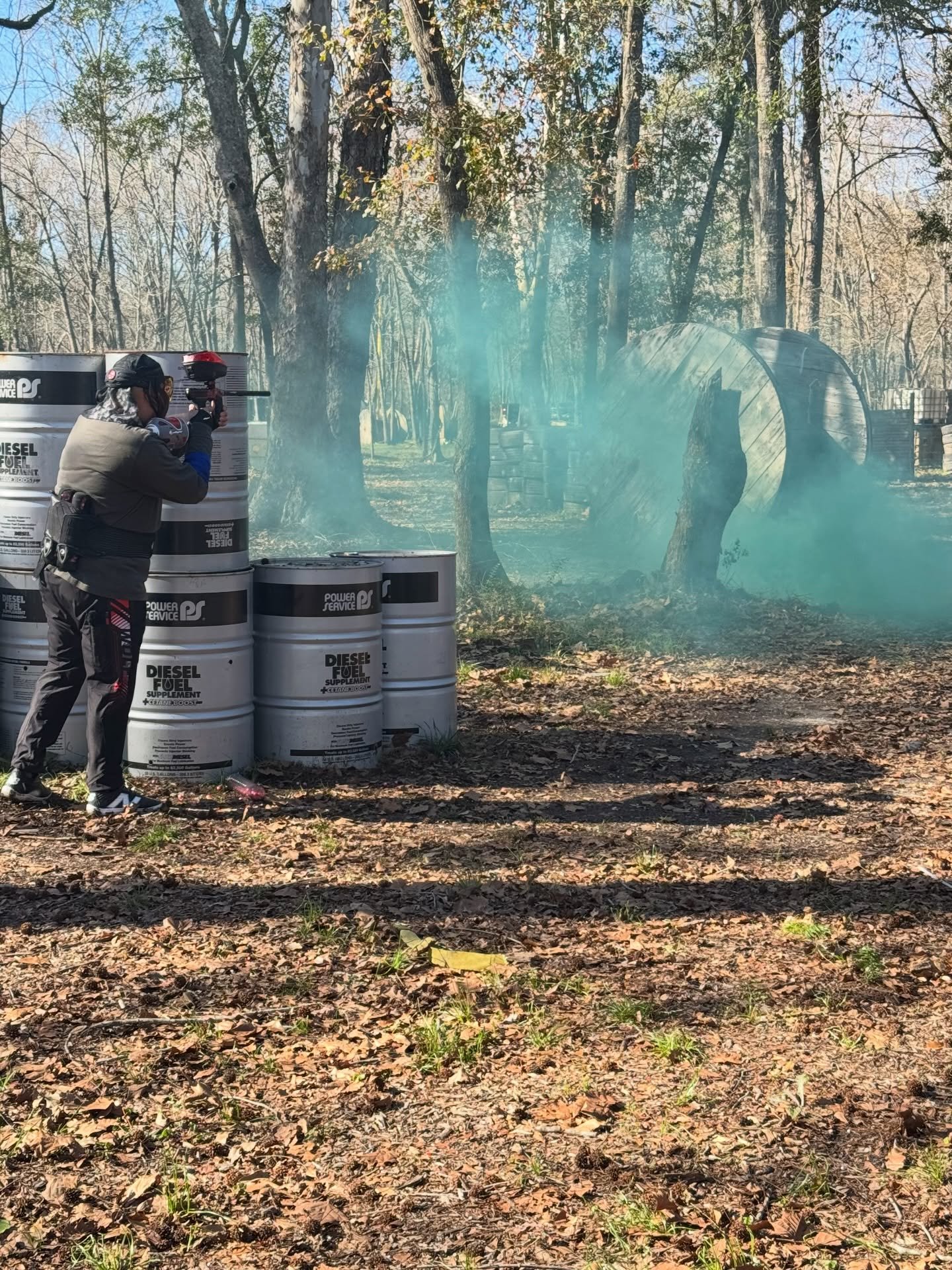 A person wearing a mask and gloves is aiming a paintball or airsoft gun behind stacked barrels in a wooded outdoor area. Smoke or fog is seen in the background amidst trees, with some barrels and a large curved metal structure also visible.