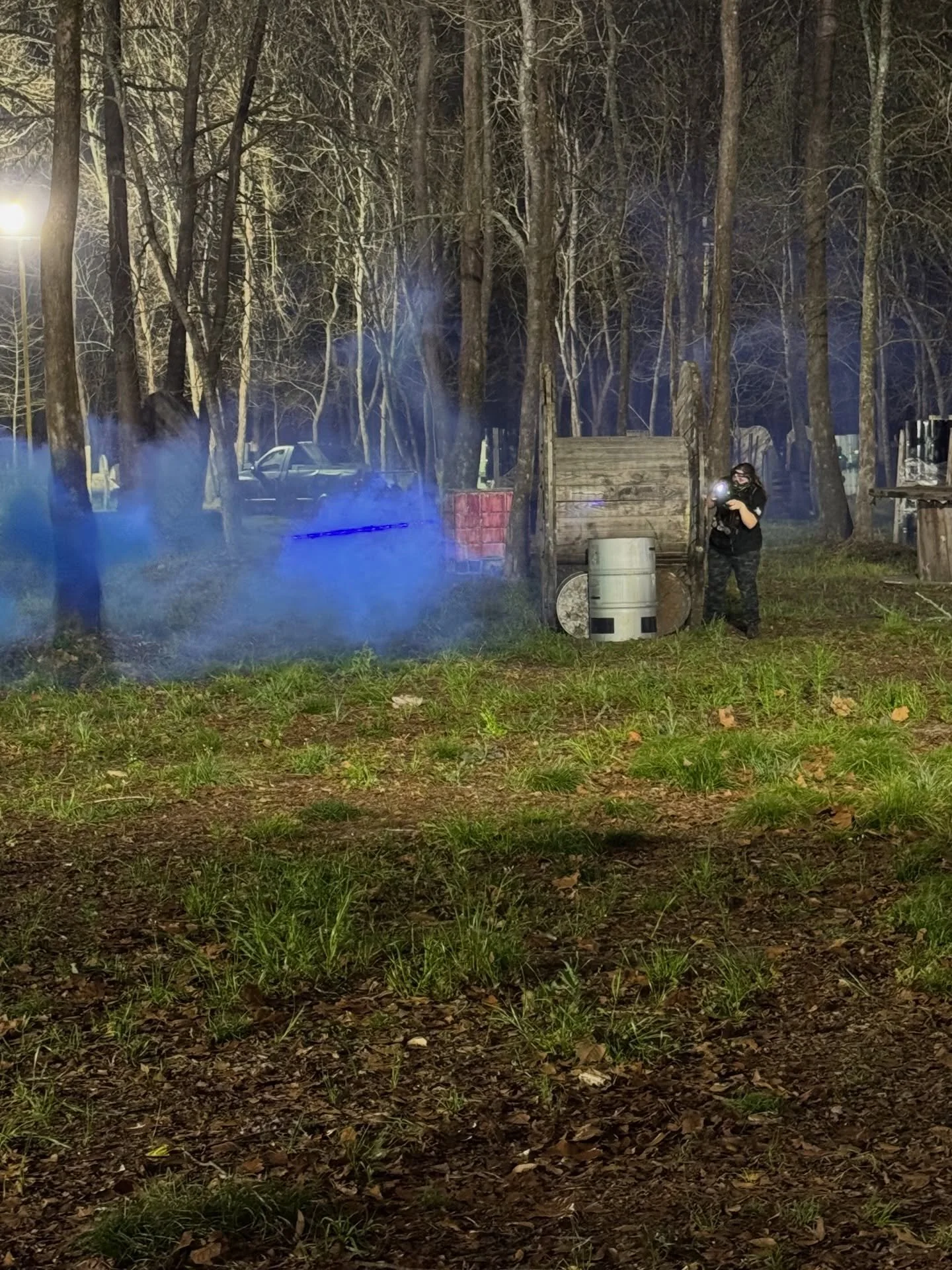 A person wearing black clothing and a mask stands in a wooded area at night, holding a flashlight, among blue smoke and wooden structures, with a parked car visible in the background.