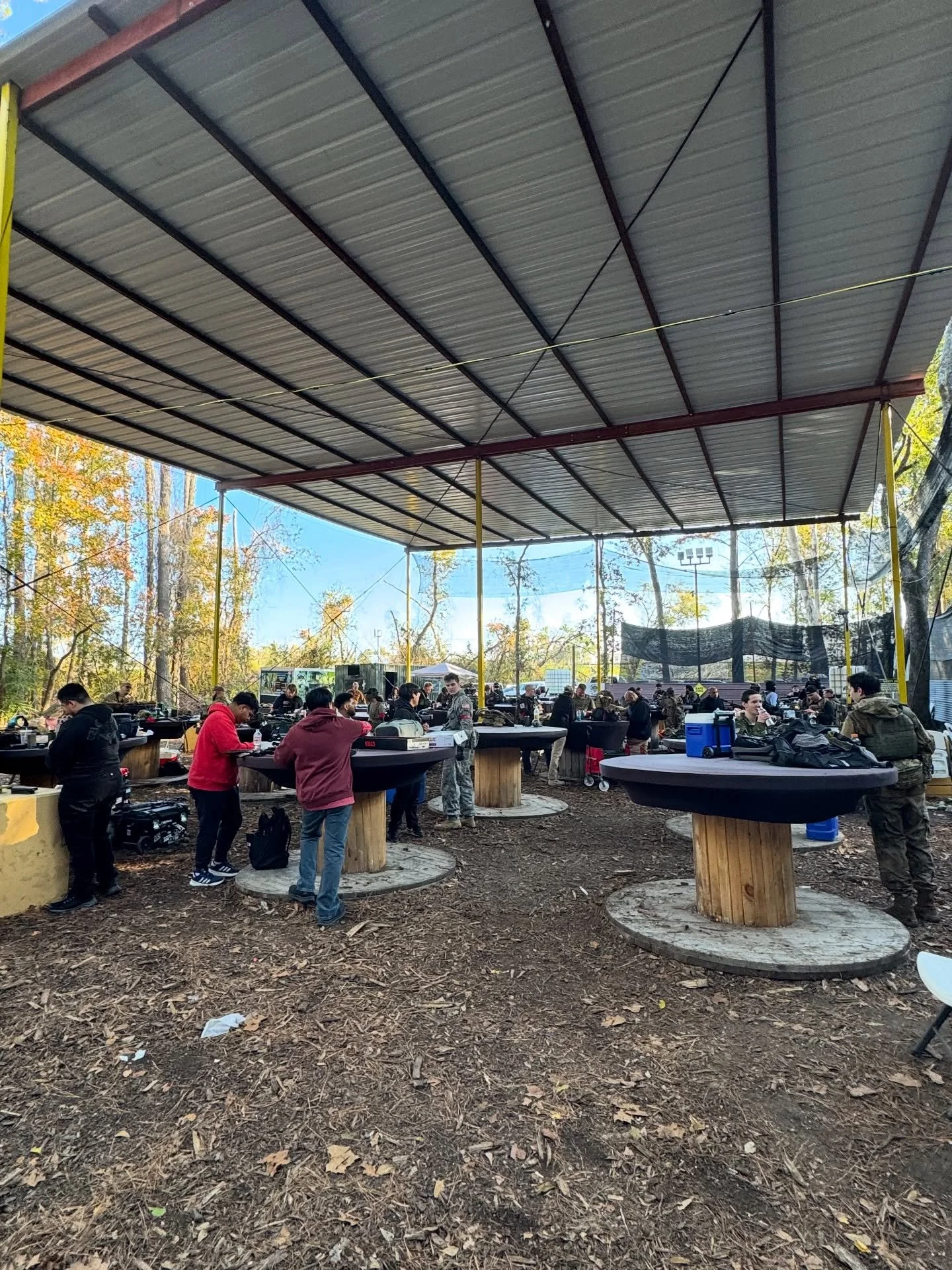 People gathered under a large outdoor canopy with tables for a group activity, with trees and a blue sky in the background.