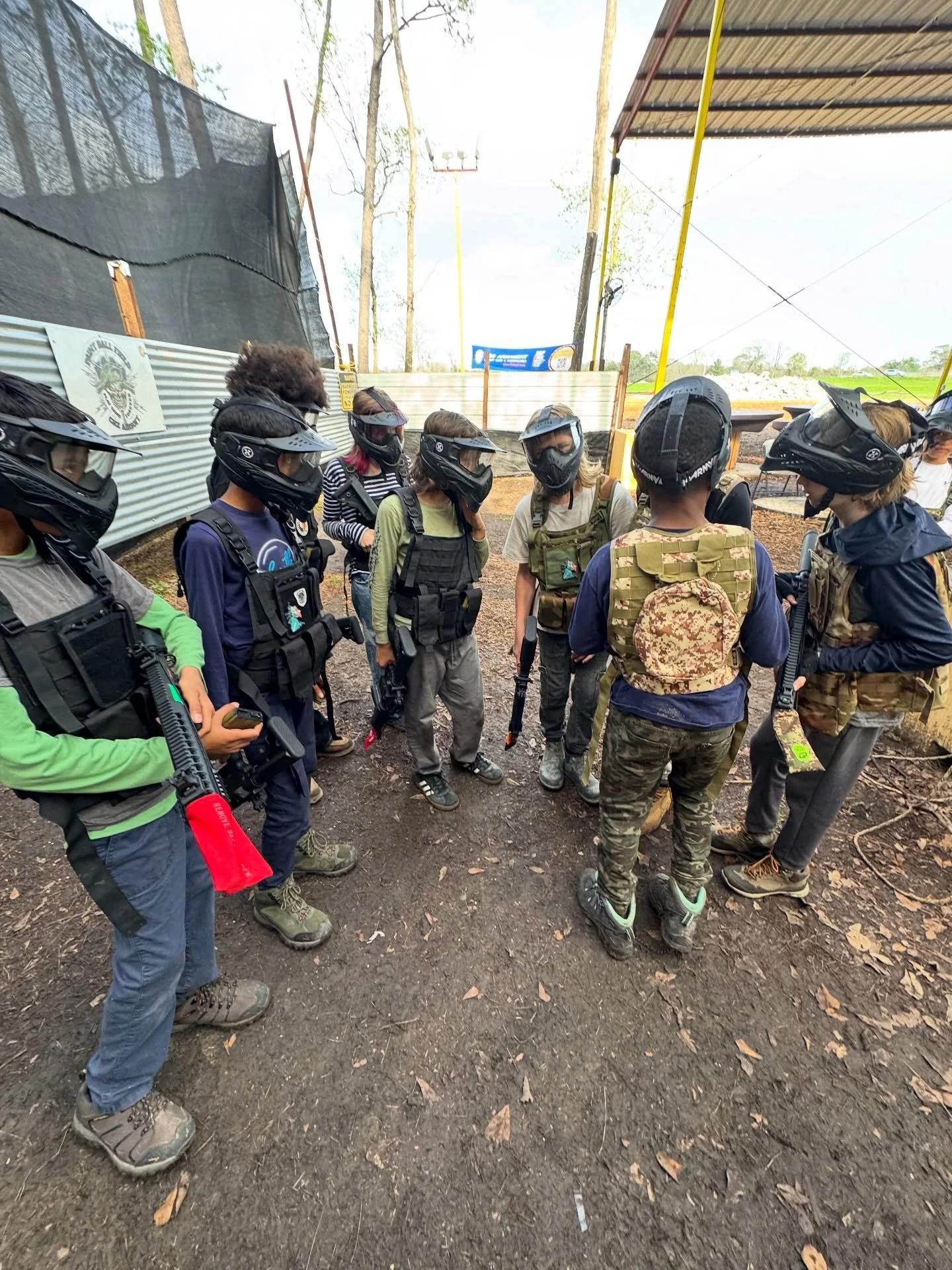 A group of people wearing paintball gear and helmets, gathered in a circle outdoors on a dirt surface.