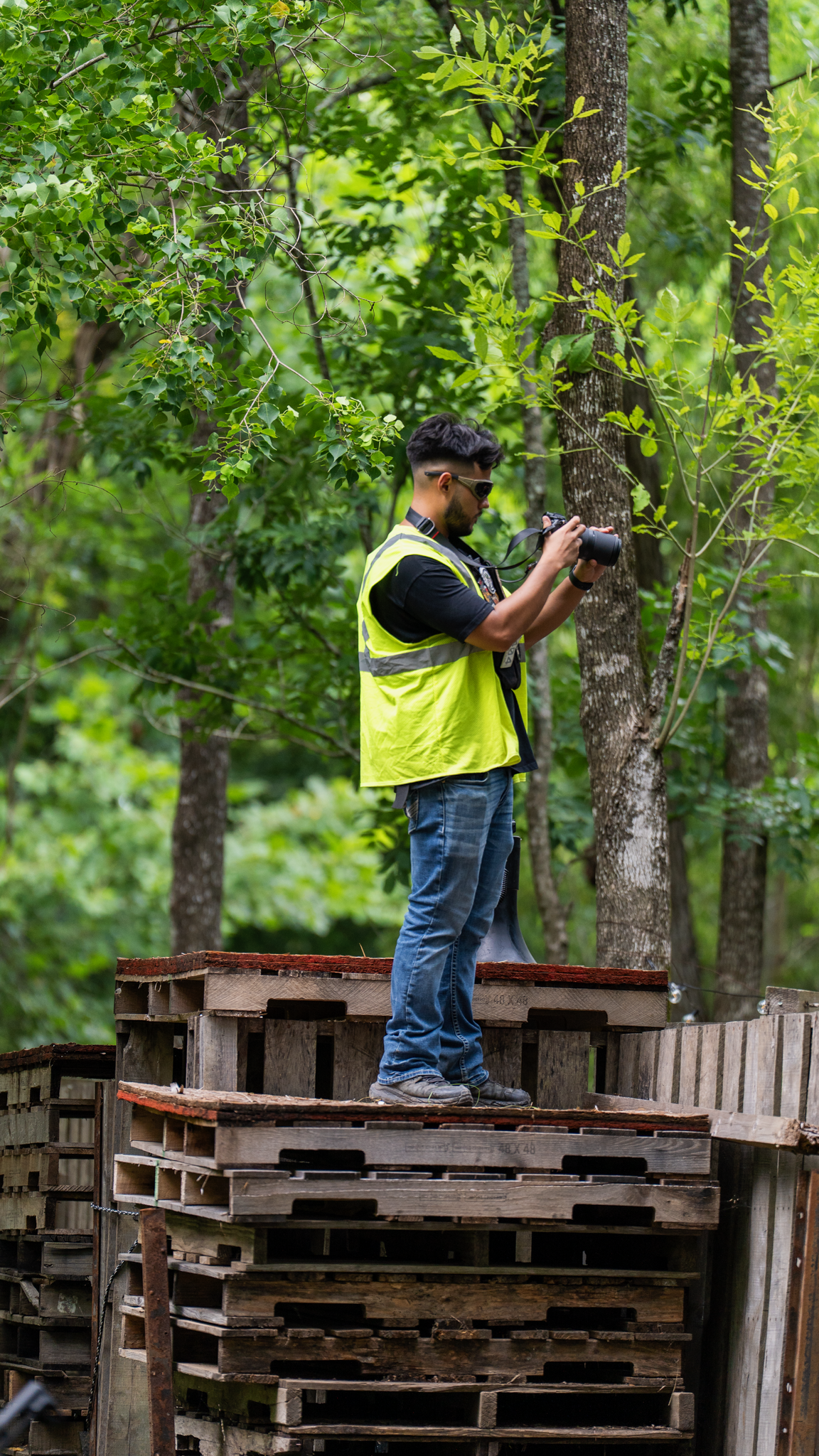 A man wearing safety goggles and a yellow safety vest stands on a stack of wooden pallets in a forest, holding a camera and looking at it.