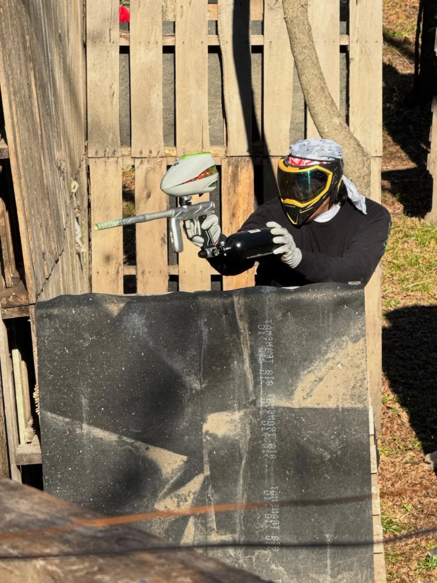 Person wearing a helmet and gloves, holding a paintball gun in a fenced outdoor area.