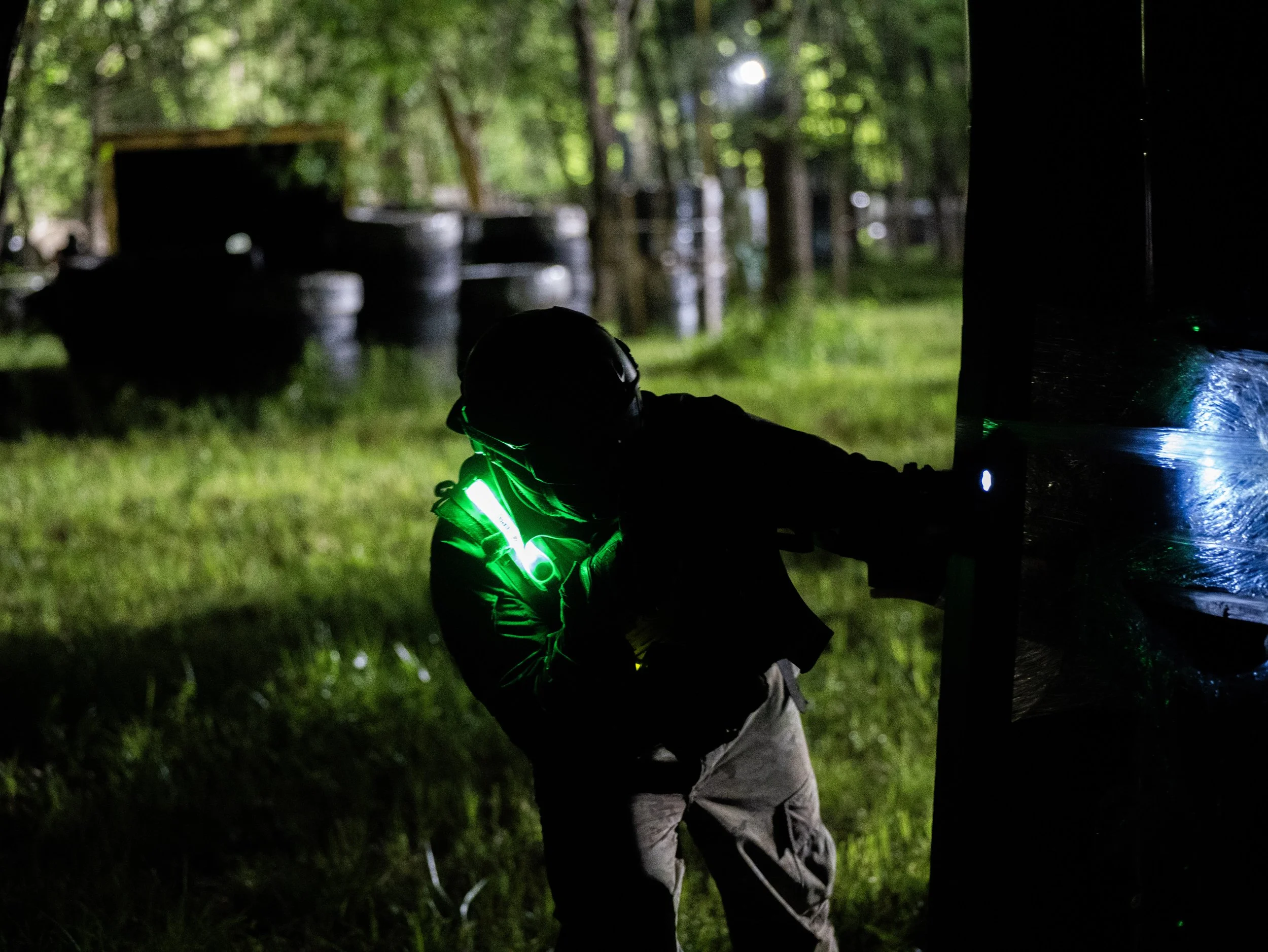 A person with a green light on their jacket is working outdoors in a wooded area at night, crouched near a dark structure, with tires and trees in the background.