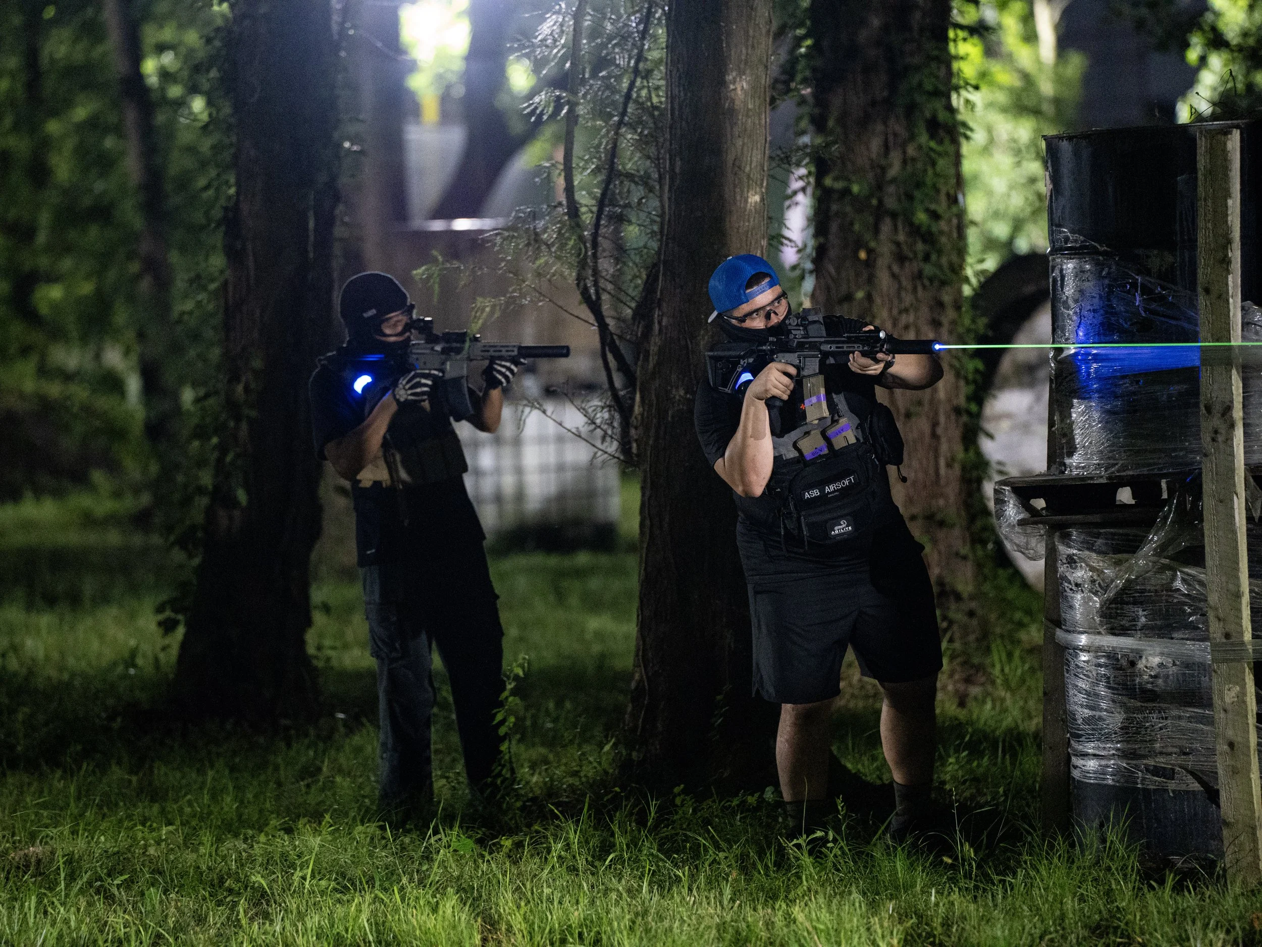 Two individuals participating in a laser tag game outdoors at night in a wooded area. They are aiming laser guns at a target on a wooden structure wrapped with plastic, with a blue laser beam visible. Both are wearing tactical vests with glow-in-the-