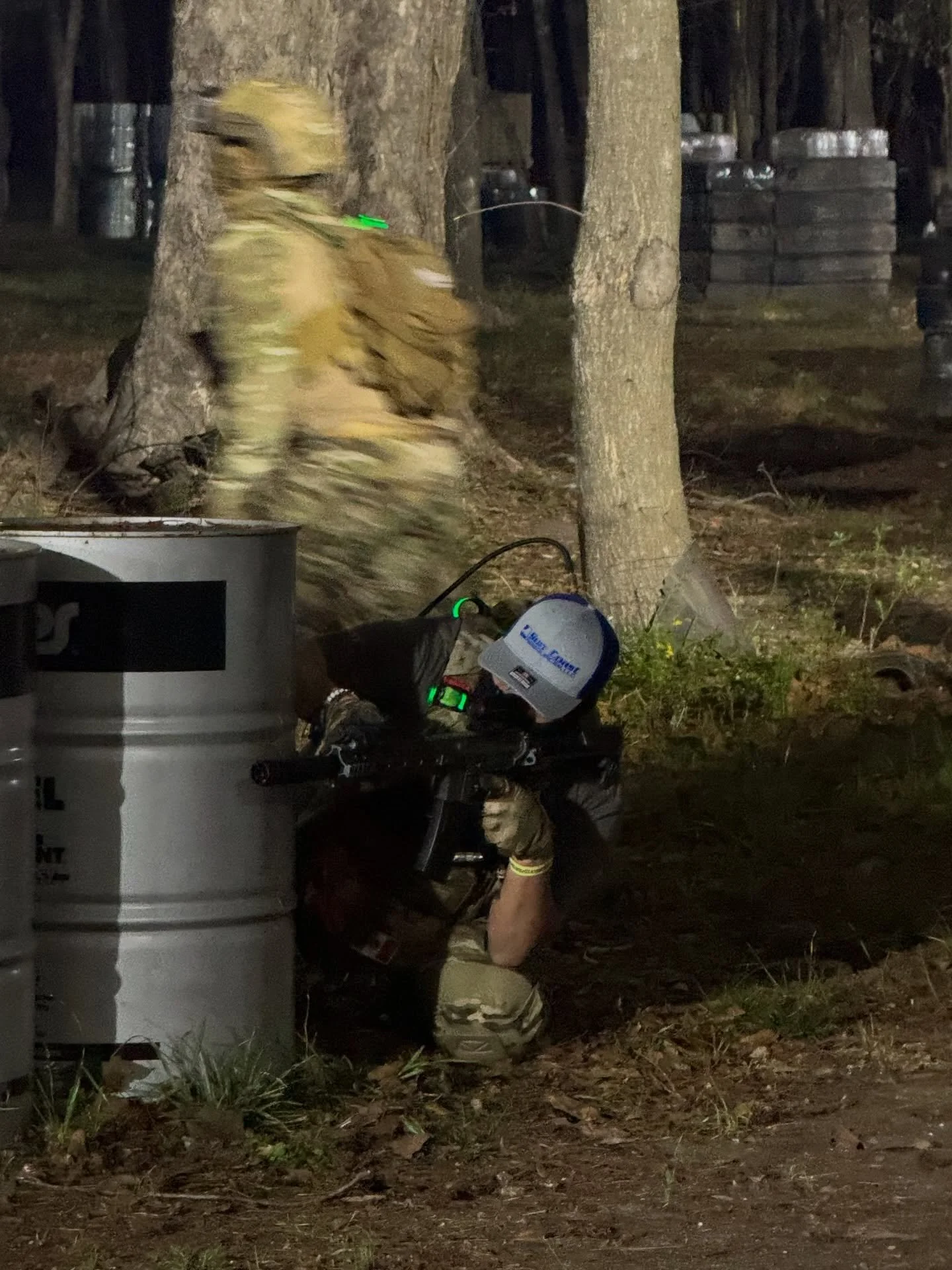 A person crouching behind a trash can with a rifle, wearing tactical gear and a helmet, in a forested area at night.