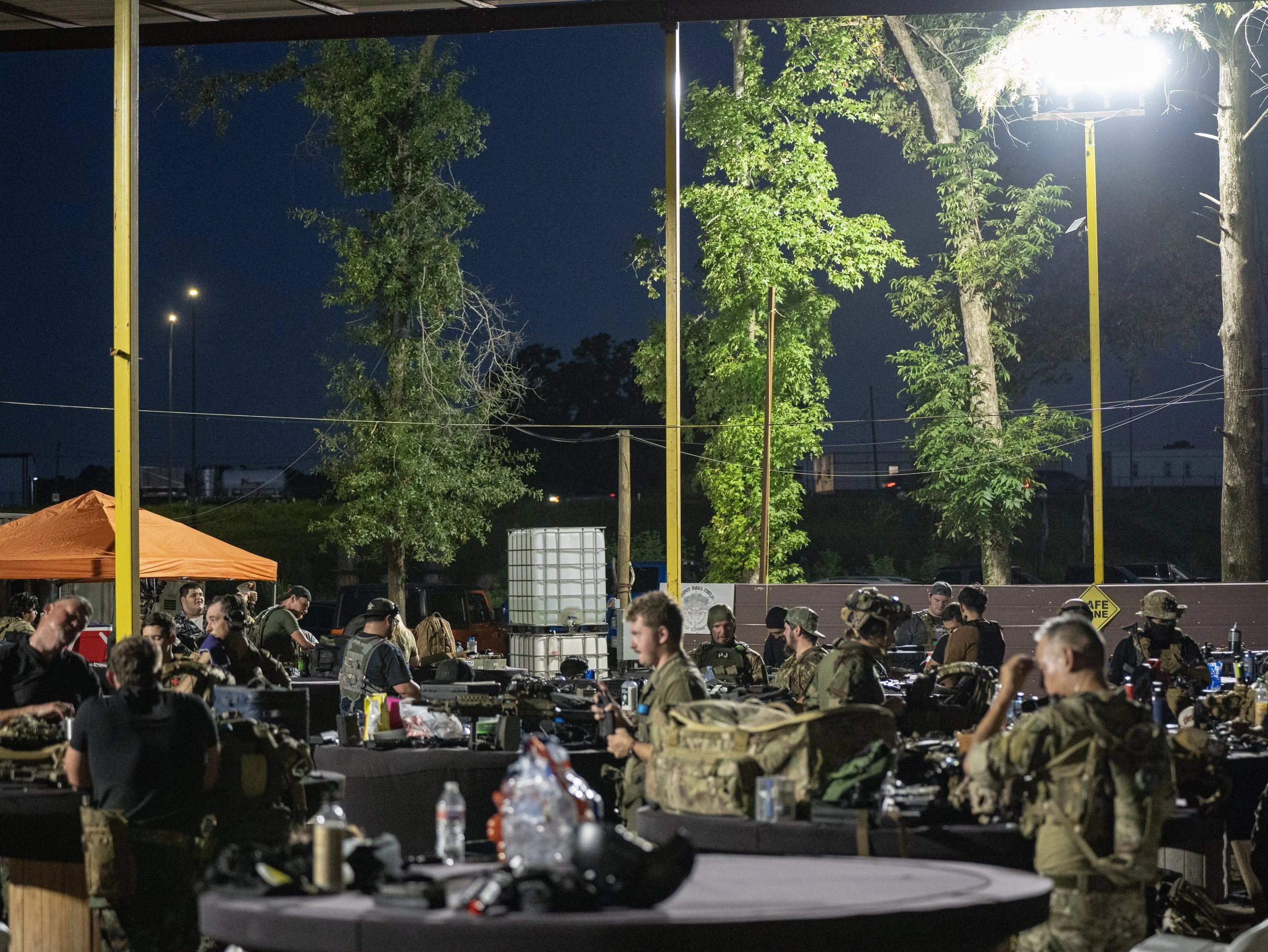 Night scene of an outdoor event with military personnel, tables, and equipment under a lit canopy, with trees and streetlights in the background.