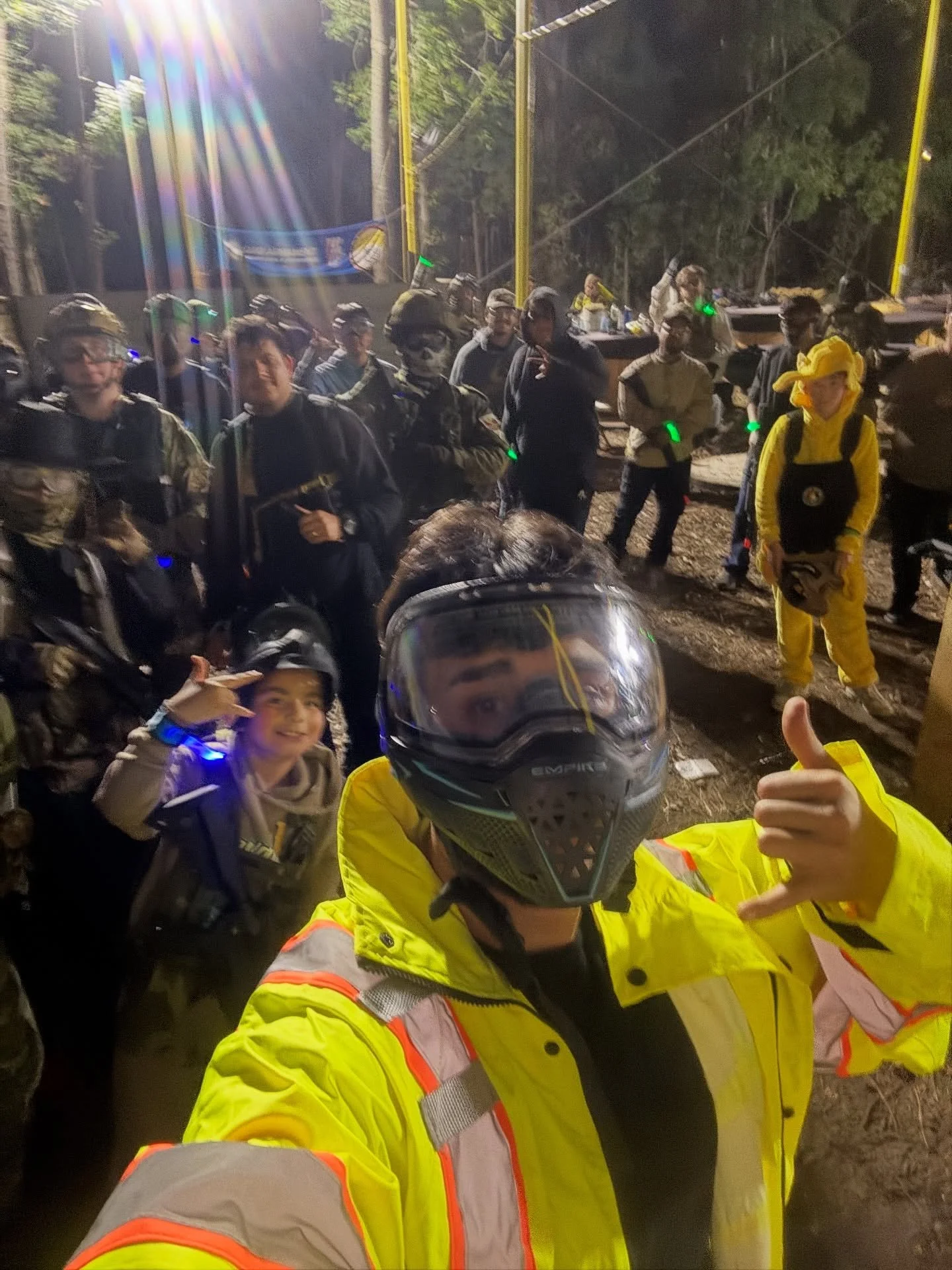 Group of people, some in paintball gear, gathered outdoors at night under lights, with trees in the background. Person in the foreground wearing a yellow jacket and helmet taking a selfie, smiling and making a hand gesture.