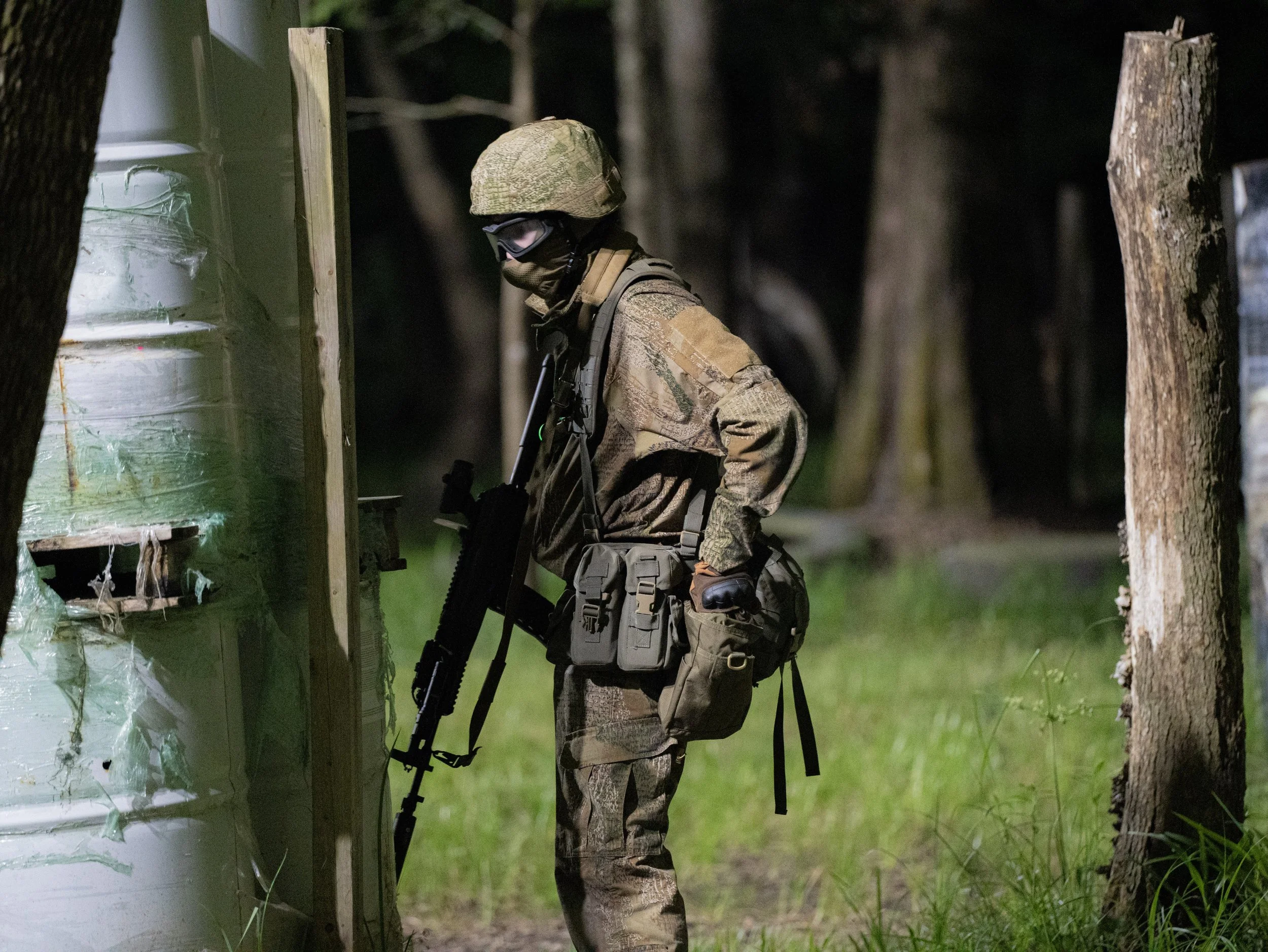 A soldier in camouflage uniform, wearing a helmet and a mask, stands in a forested area at night, inspecting a damaged green and white structure.