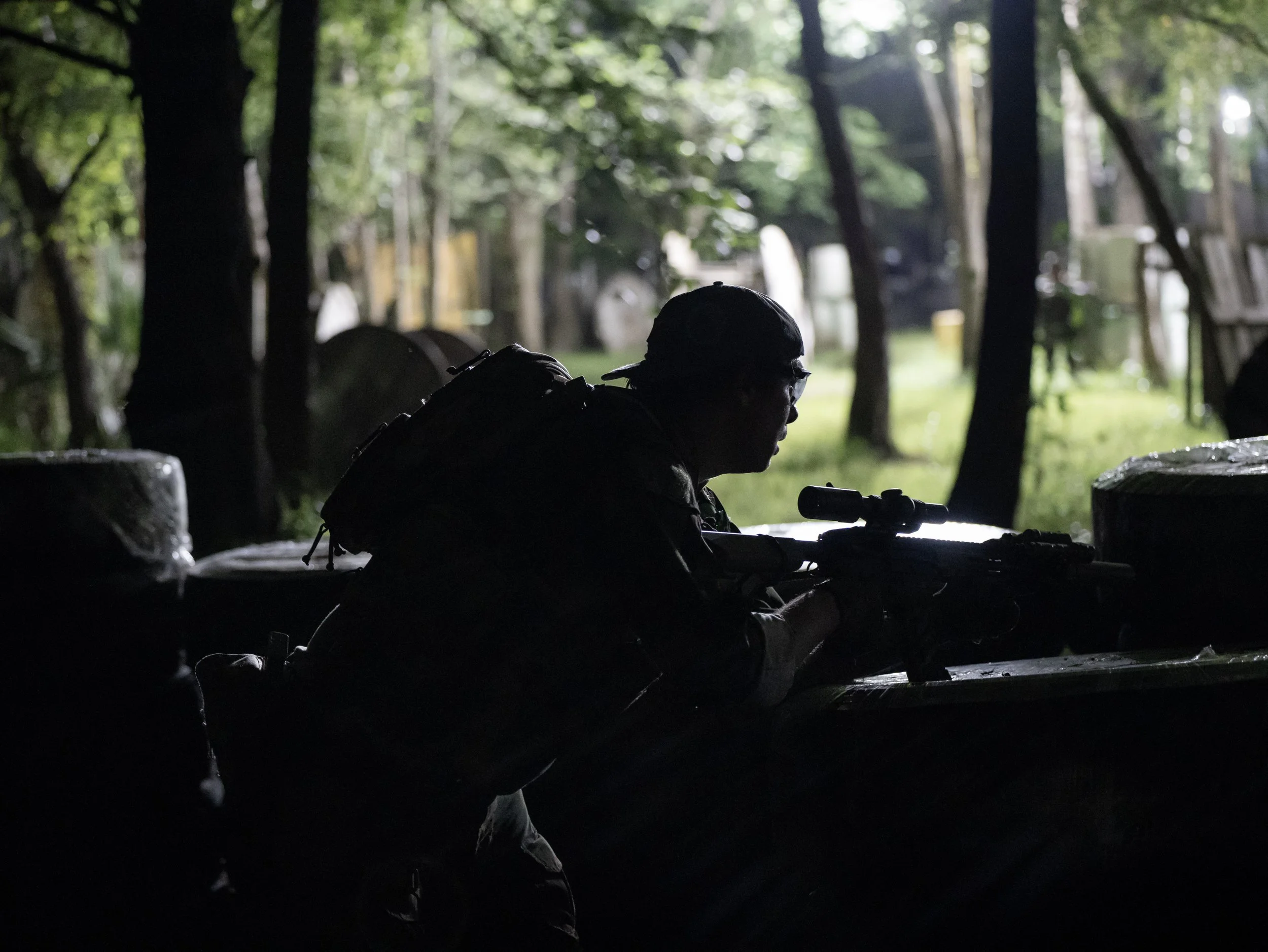 Silhouette of a soldier in military gear with helmet and binoculars in a dark forest with trees and greenery in the background.