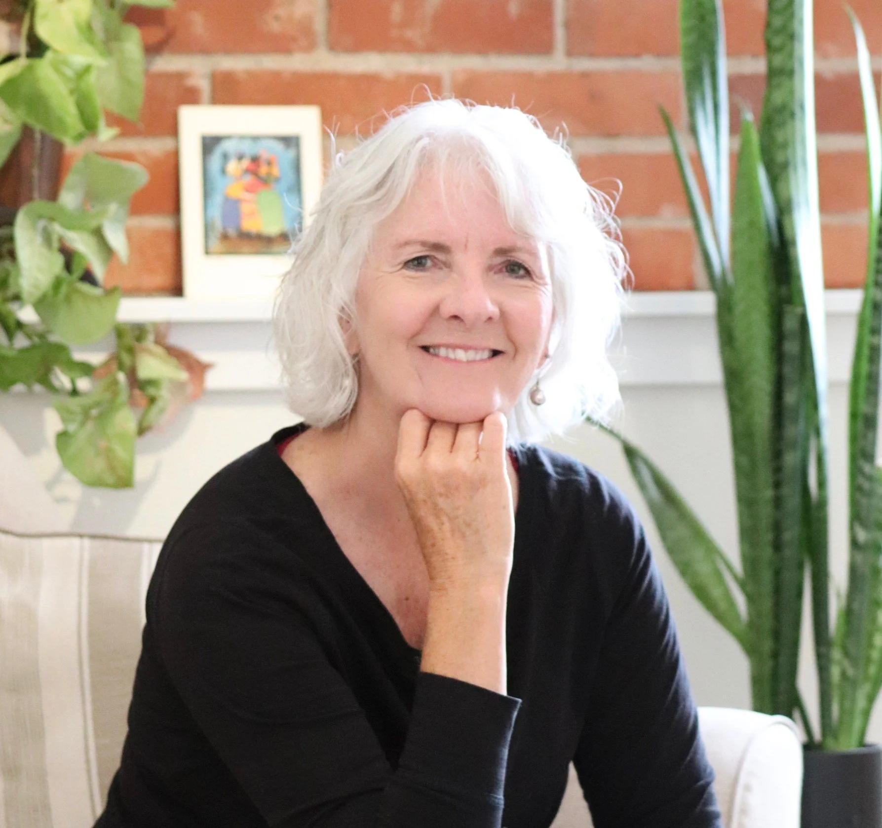 A smiling elderly woman with white hair, wearing a black top, sitting indoors surrounded by green plants and a framed picture on a red brick wall in the background.