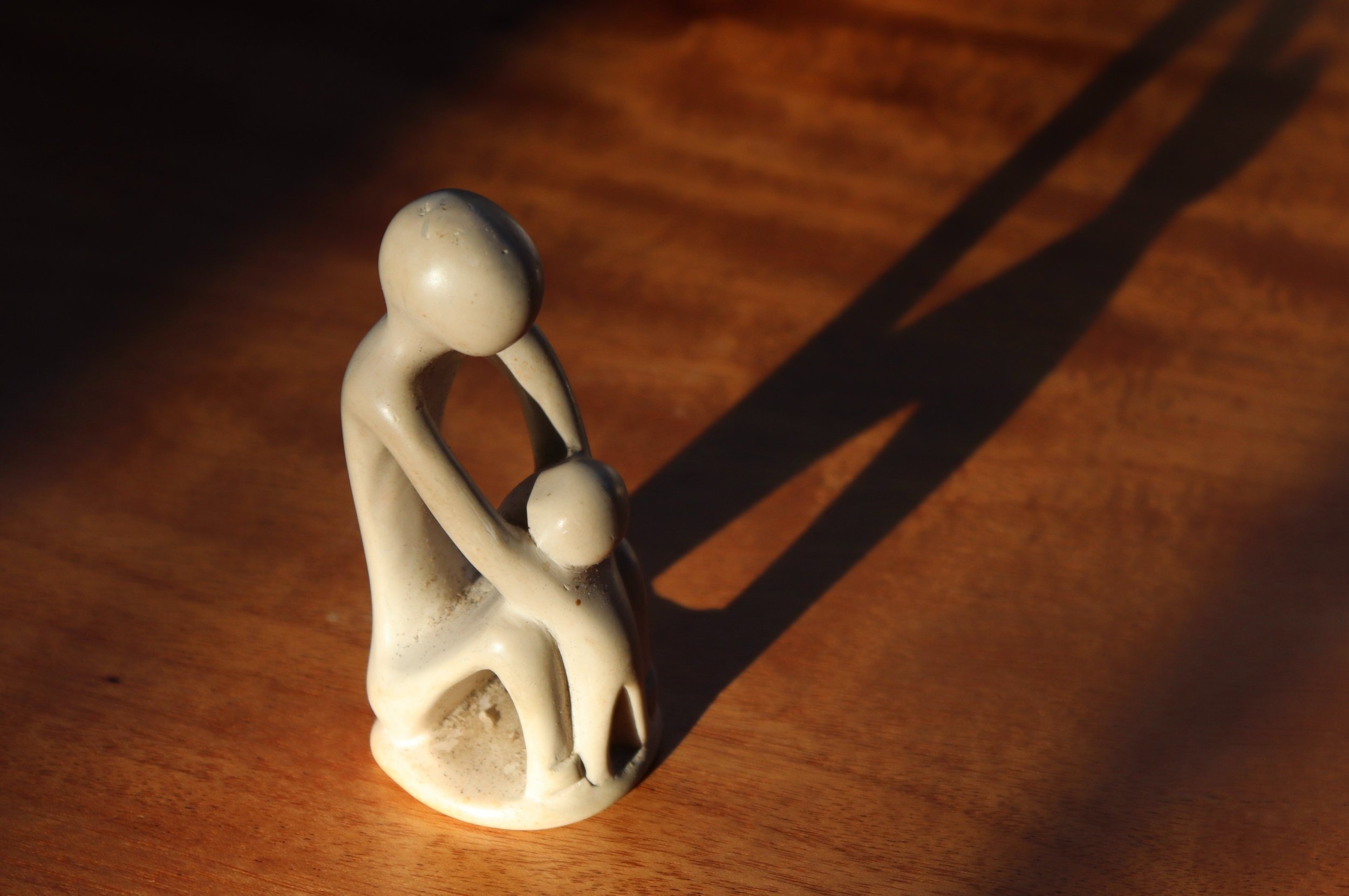 A small sculpture of a human figure with its head bowed and hands covering its face, placed on a wooden surface. The sculpture is white and casts a shadow on the wood.