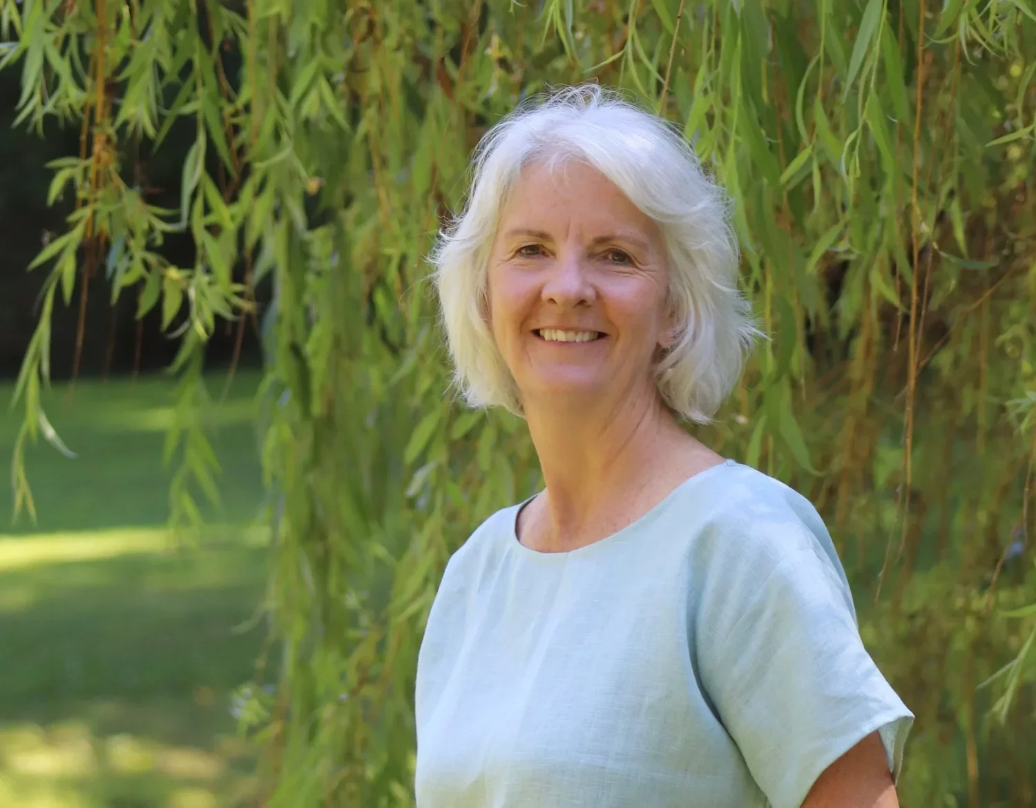 A smiling elderly woman with white hair standing outdoors near a tree with hanging branches and leaves.