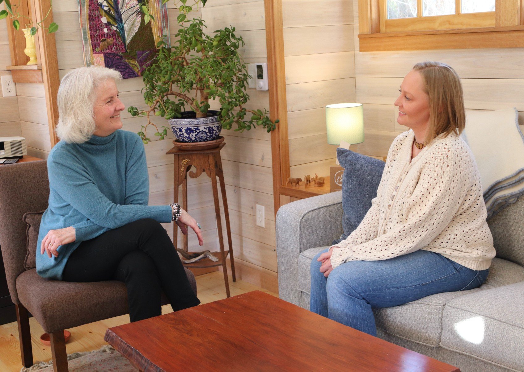 Two women sitting and talking in a cozy living room with wooden walls. One woman has gray hair, wears a blue turtleneck and black pants, while the other has blonde hair, wears a cream sweater and blue jeans. There is a large green plant behind them and a warm lamp on a side table.