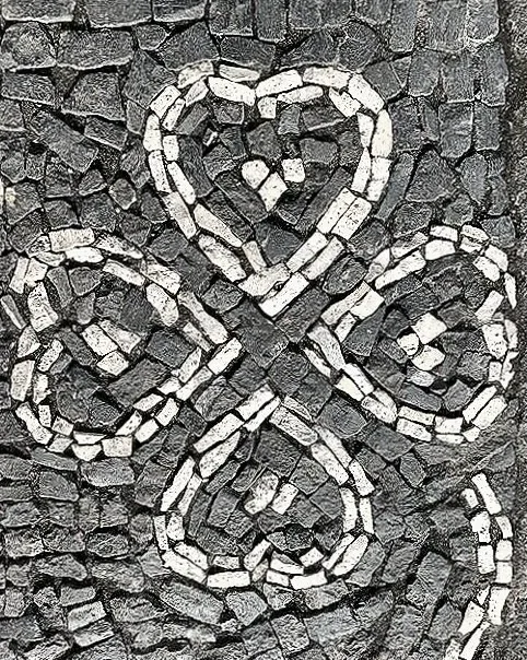 A mosaic of stones arranged in the shape of a four-leaf clover with a heart at the top center, set against a background of dark, irregularly shaped stones.