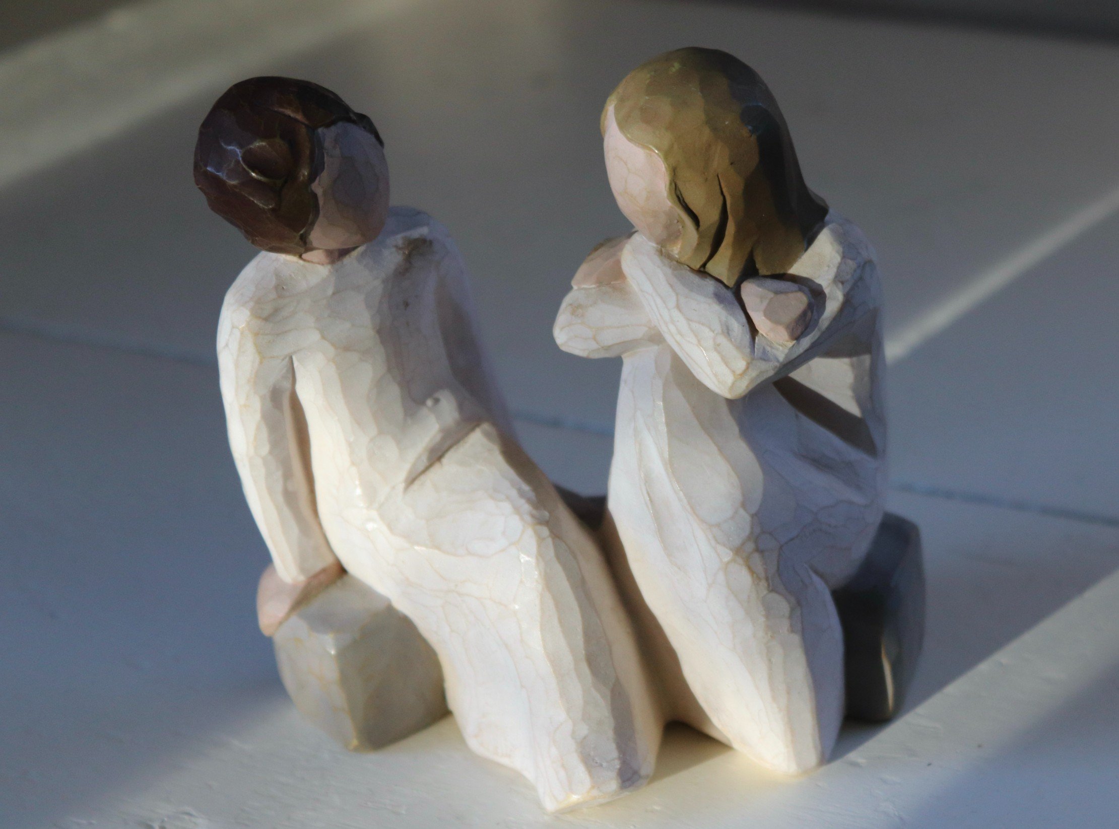 Two small figurines made of carved stone or marble, resembling ancient sculptures, depict two people with shaved heads, one with darker hair and the other with lighter hair, sitting in a contemplative posture.