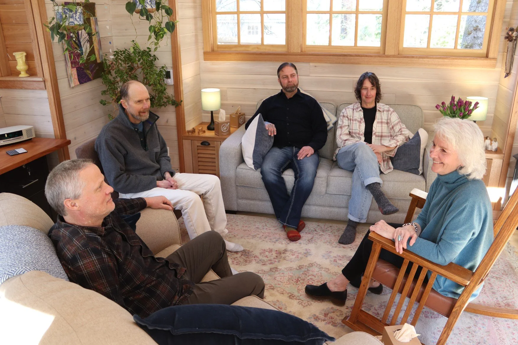 Six people sitting in a cozy living room with wooden walls and large windows, engaged in conversation and smiling.