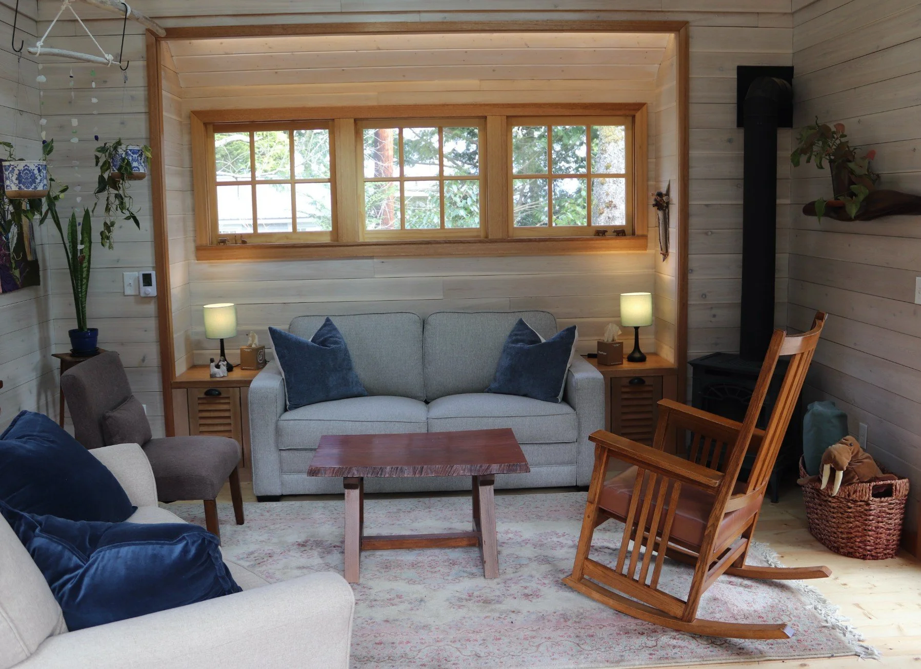 Living room with wooden walls, large window, sofa, armchairs, wooden rocking chair, area rug, potted plant, lamps, and decorative items.
