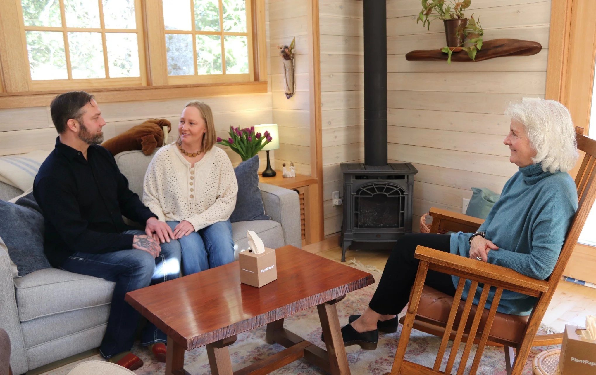 Four people sitting in a cozy living room having a conversation, with a wood stove, a table with tissues, and a window with trees outside.