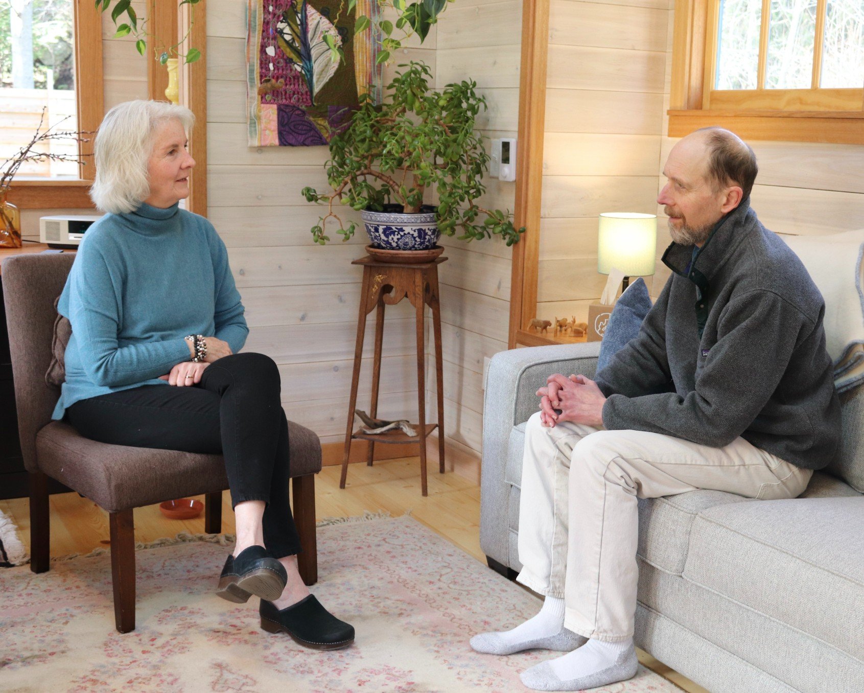 An elderly woman and a middle-aged man are sitting indoors facing each other, engaged in conversation. The woman has white hair and is wearing a blue turtleneck sweater and black pants. The man has a beard and is dressed in a gray jacket with white pants and white socks. The room features lightwood-paneled walls, a window, and decorative items including a large potted plant, artwork, and a small lamp.