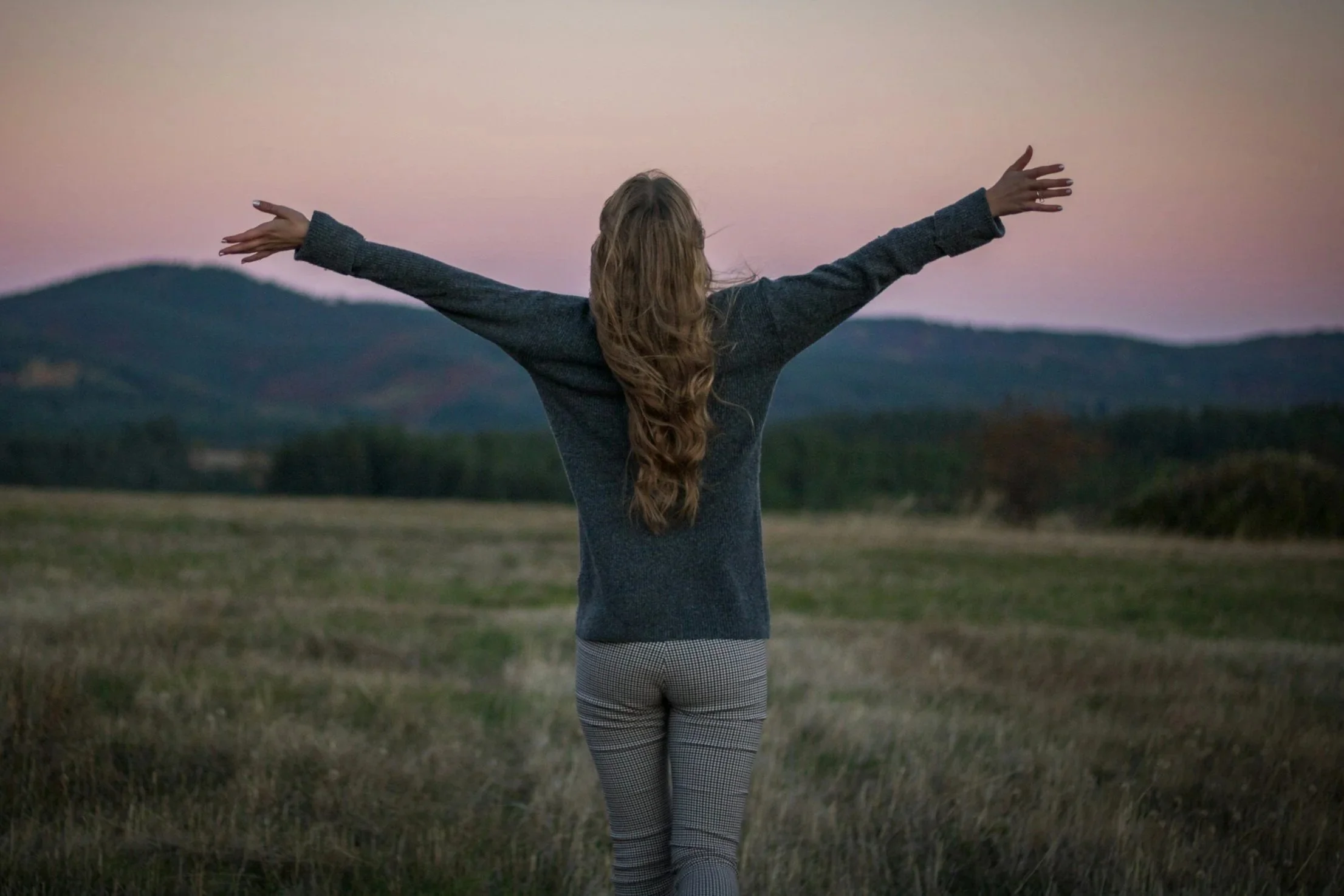 Woman walking outdoors with arms outstretched at sunset or dusk, with mountains in the background.