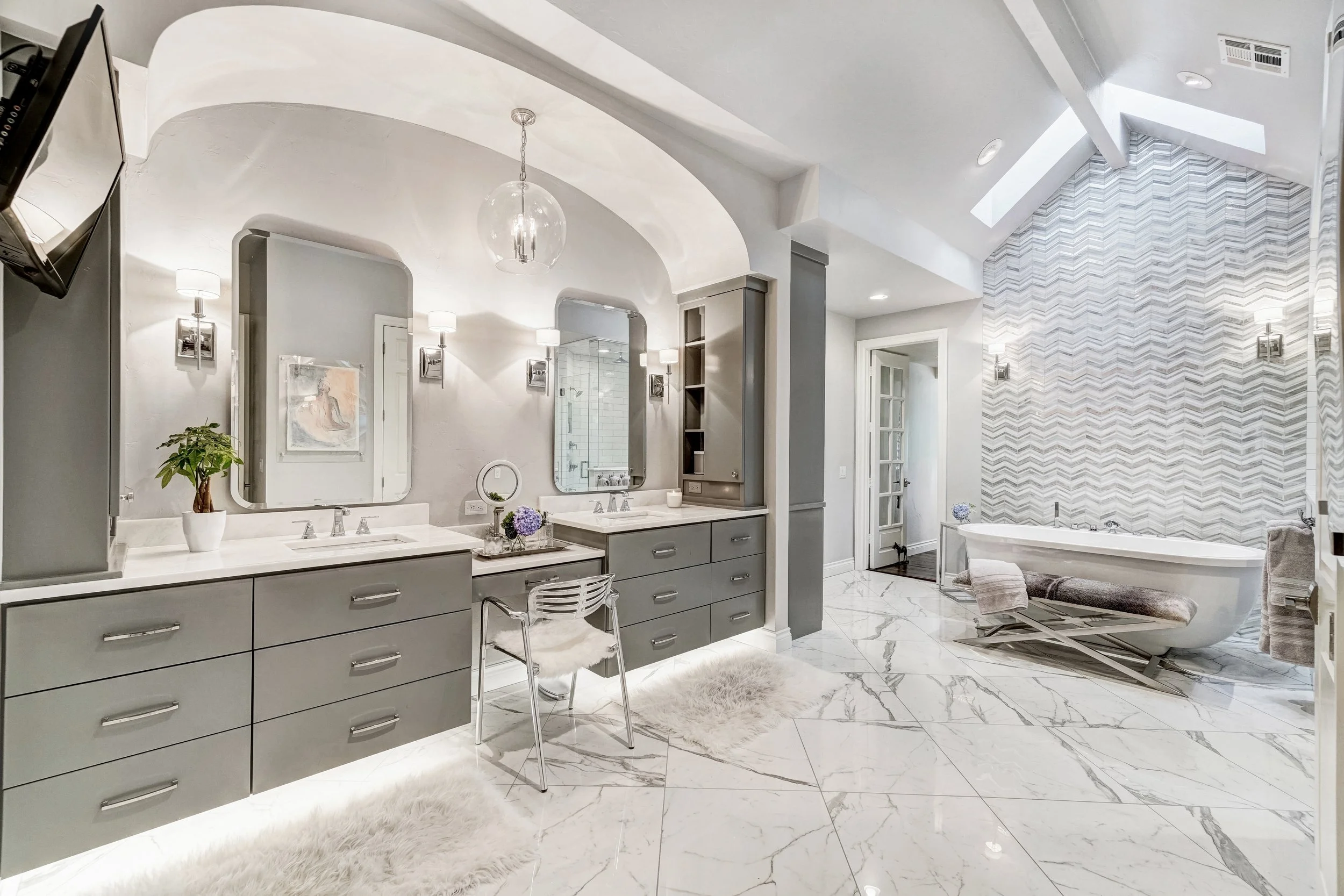After renovation: Double vanity with gray cabinetry, quartz countertops, framed mirrors, makeup station, arched ceiling detail, freestanding bathtub, chevron tile feature wall, large-format marble-look flooring, and decorative wall sconces.