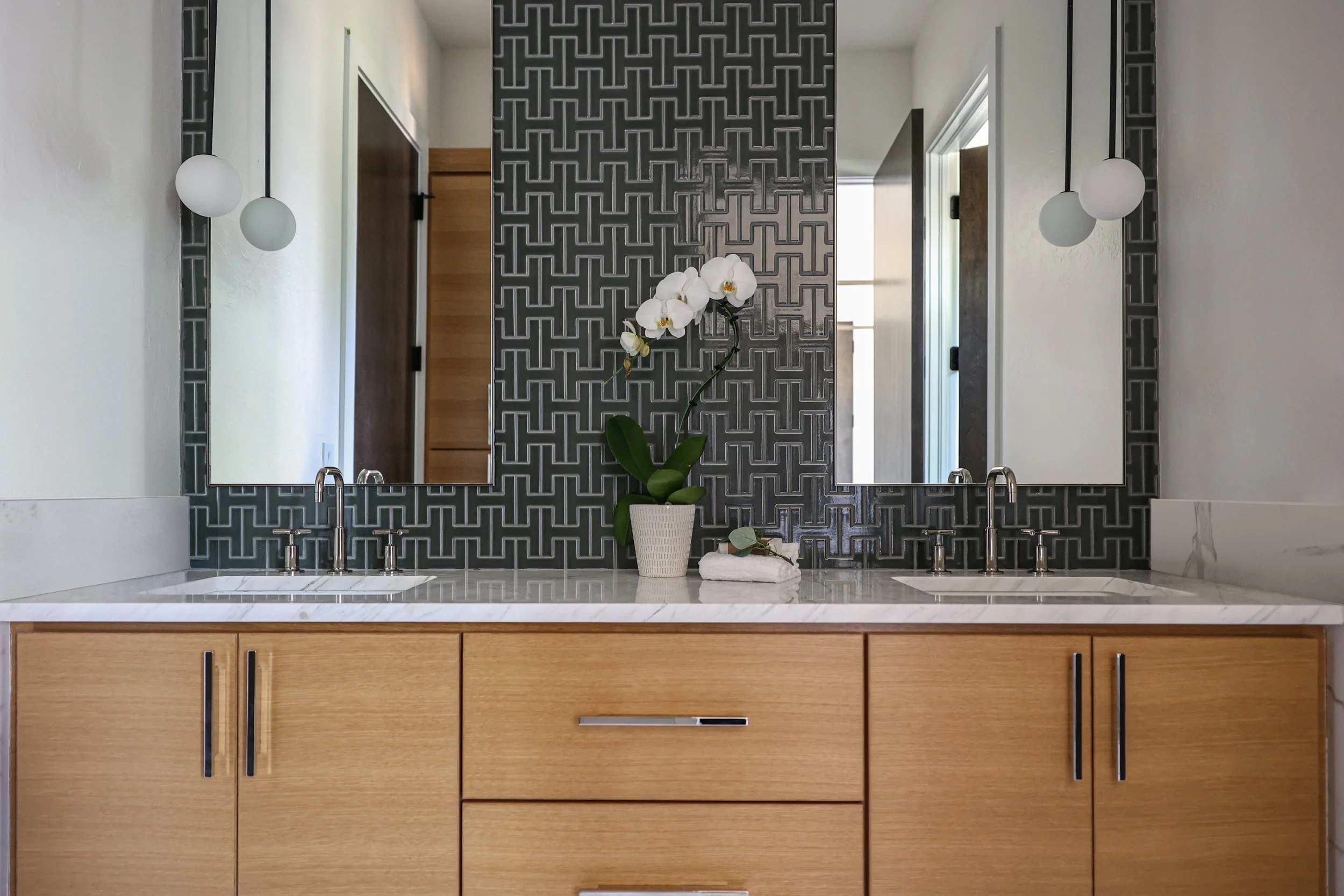 Double vanity with light wood cabinetry, white countertop, rectangular mirrors, geometric tile backsplash, chrome faucets, globe pendant lights, and orchid centerpiece.