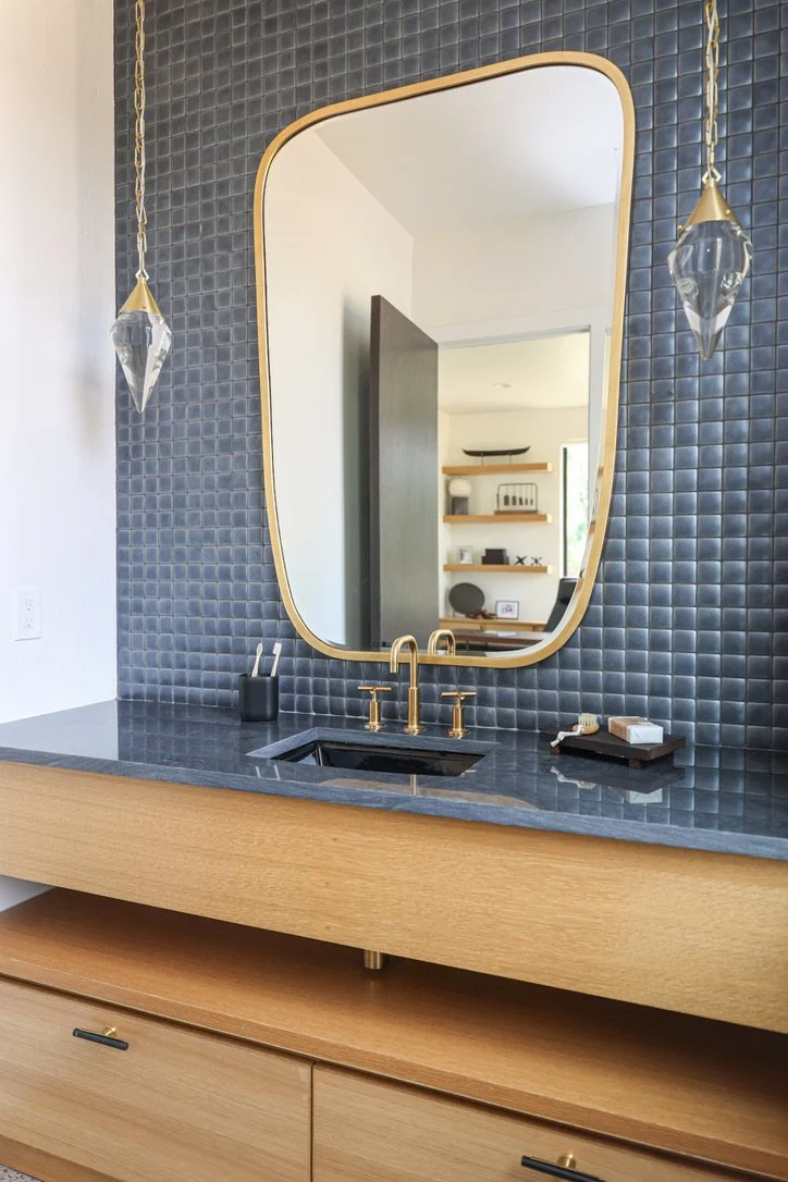 Floating wood vanity with dark countertop, black tile wall, brass faucet, asymmetrical framed mirror, pendant lights, integrated sink, and open shelving visible through doorway.