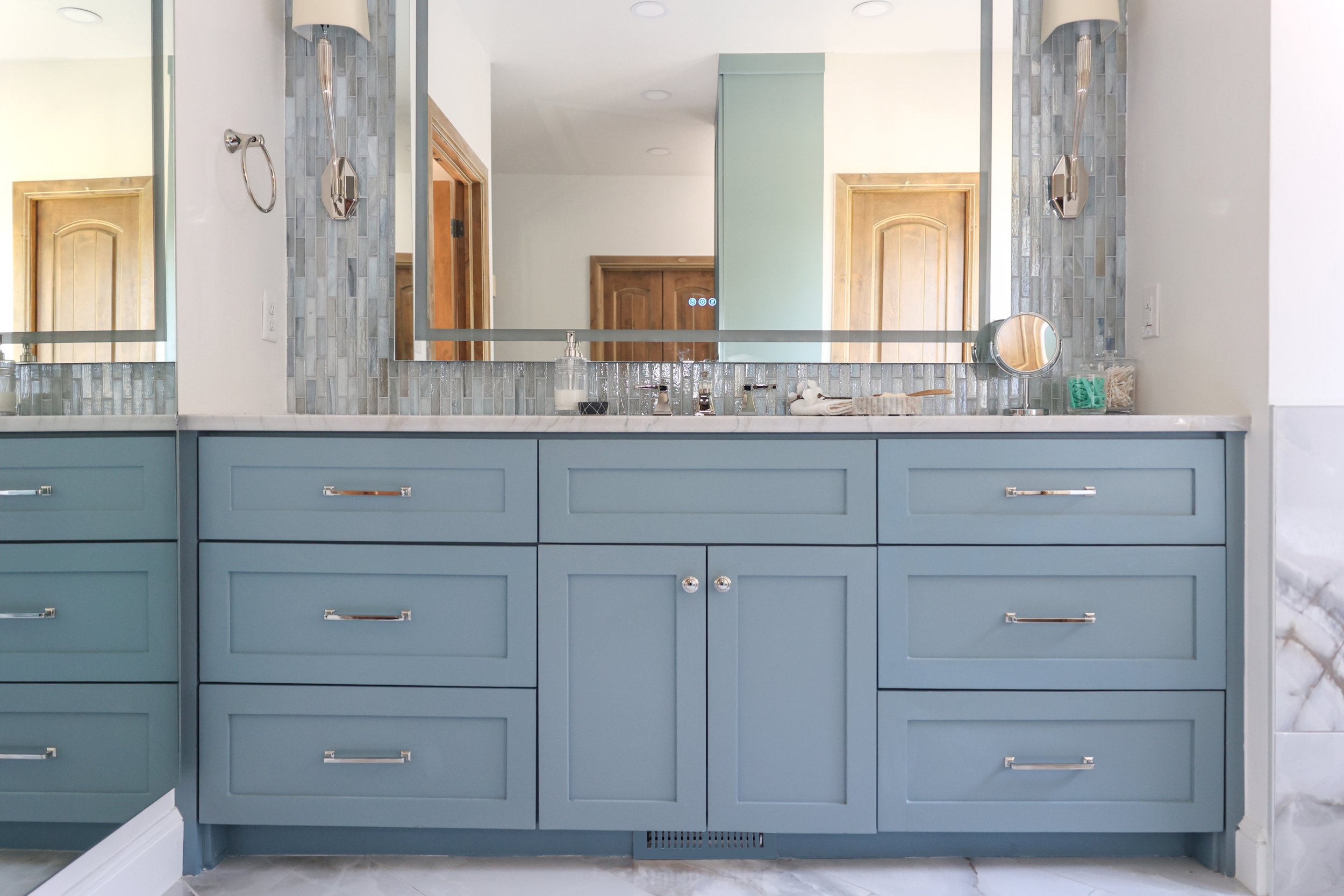 Blue double vanity with shaker-style cabinetry, marble countertop, full-width mirror, mosaic tile backsplash, wall sconces, chrome hardware, and large-format floor tile.