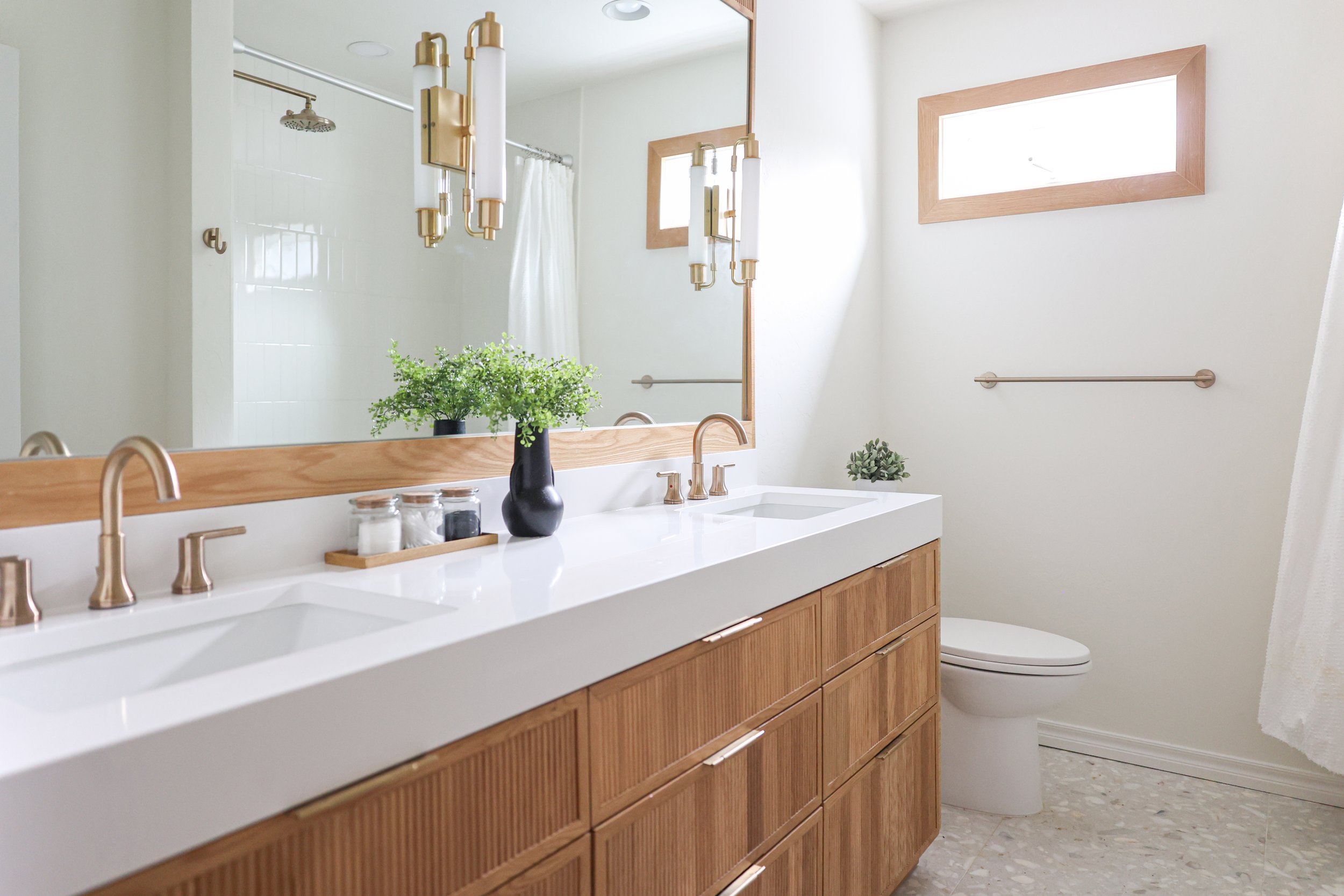 Double vanity with white integrated countertop, wood cabinetry, framed mirror, brass wall sconces, dual faucets, shower with curtain, terrazzo flooring, and clerestory window.