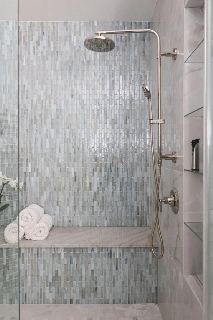 Walk-in shower with glass enclosure, marble-look wall tile, vertical mosaic tile accent wall, built-in bench, recessed shelving, rainfall showerhead, and handheld shower fixture.