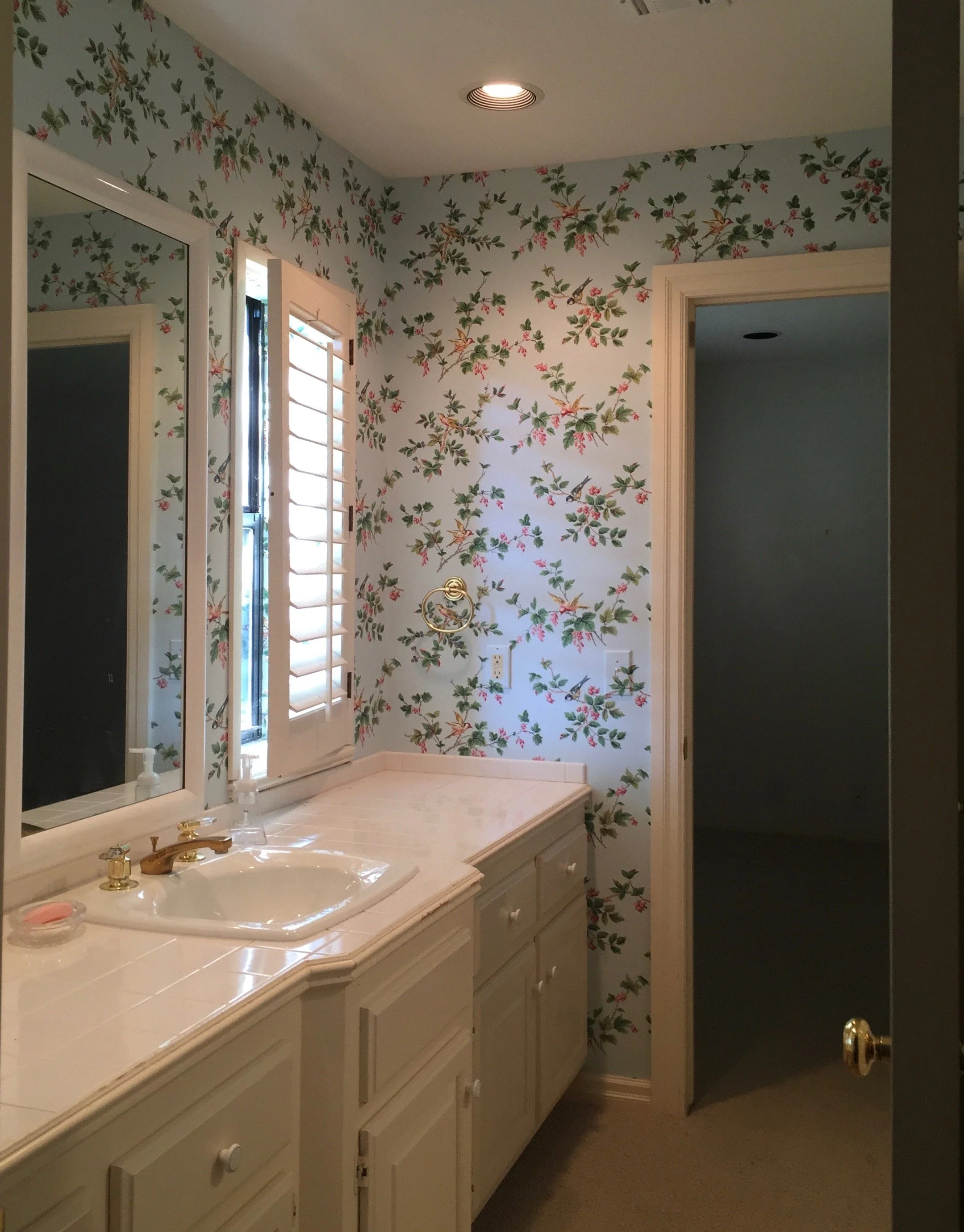Before renovation: Single vanity with tiled countertop, undermount sink, framed mirror, floral wallpaper, plantation shutters, brass faucet, recessed lighting, and painted wood cabinetry.