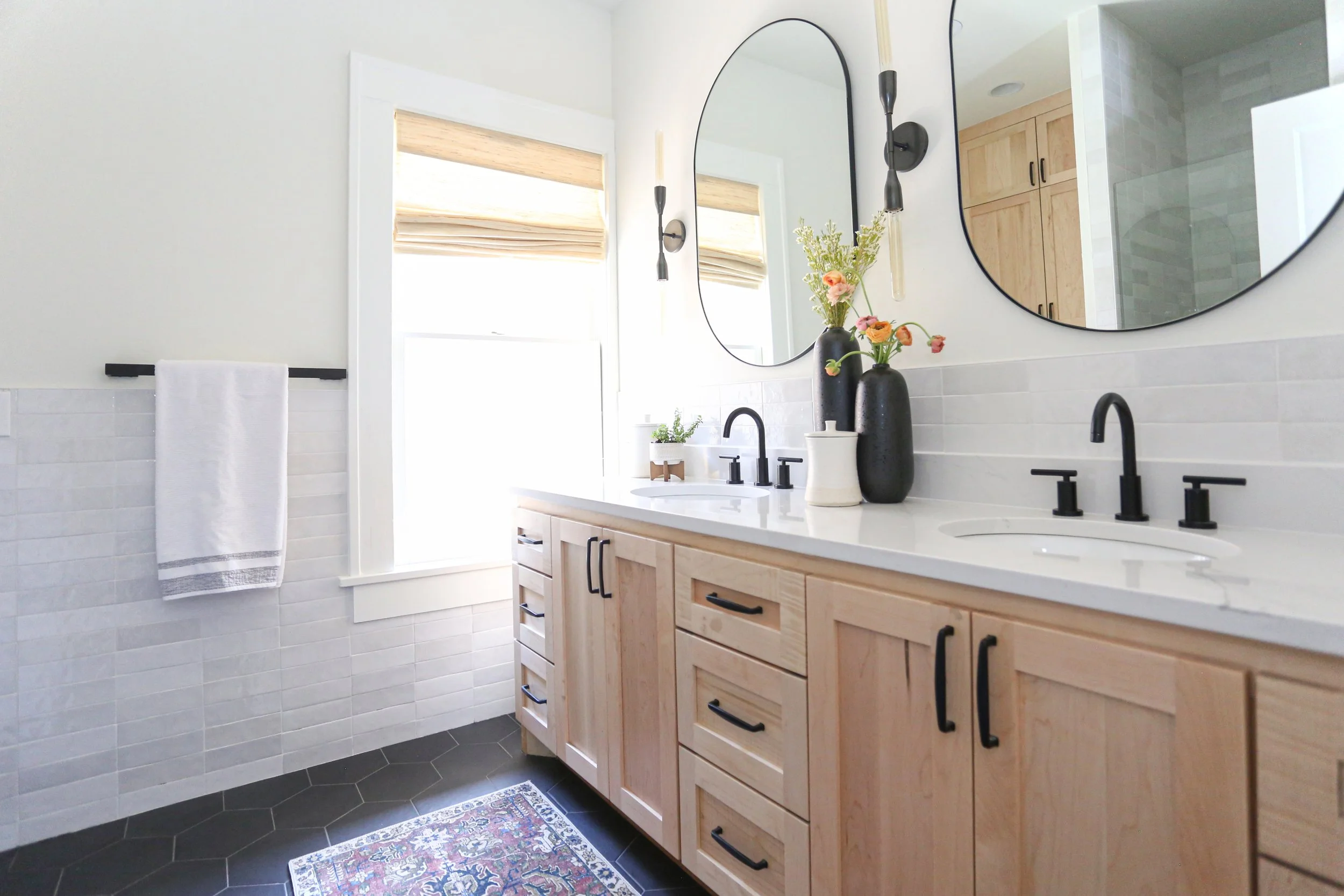 Double vanity with light wood cabinetry, white countertop, oval mirrors, black plumbing fixtures, wall sconces, subway tile wainscoting, hexagon floor tile, and window with woven shade.