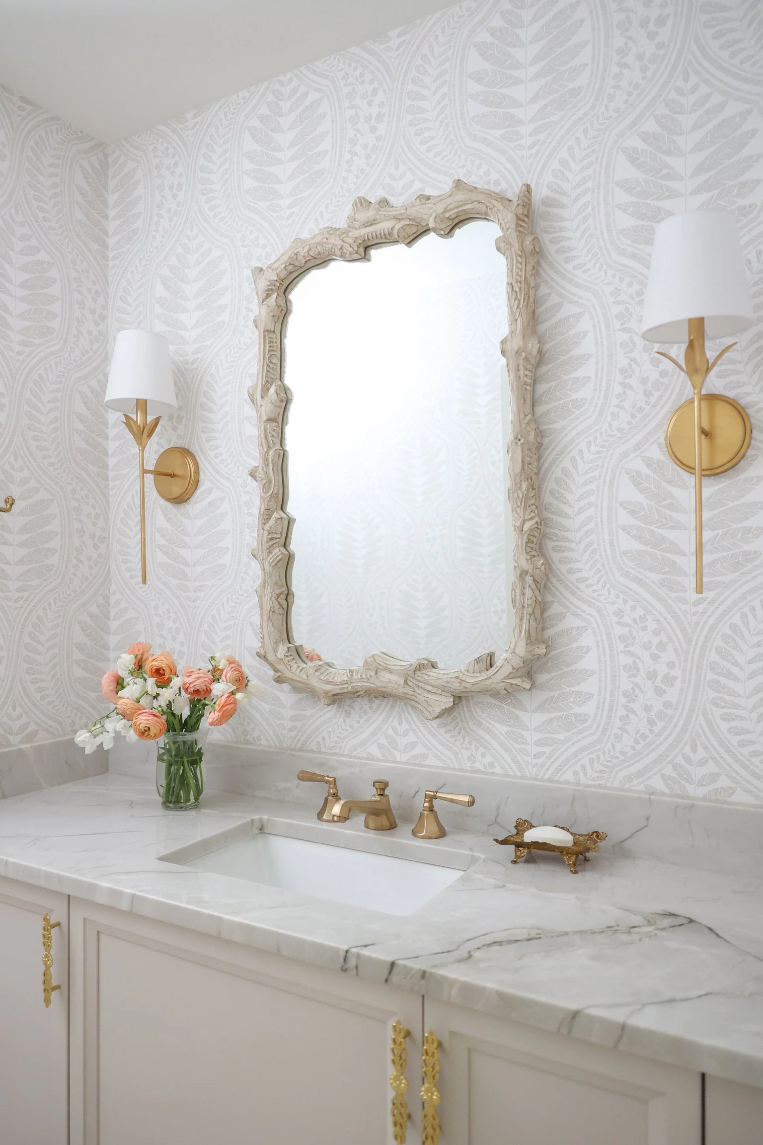 White vanity with decorative brass hardware, marble countertop, undermount sink, brass faucet, carved framed mirror, wall sconces, patterned wallpaper, and floral arrangement.