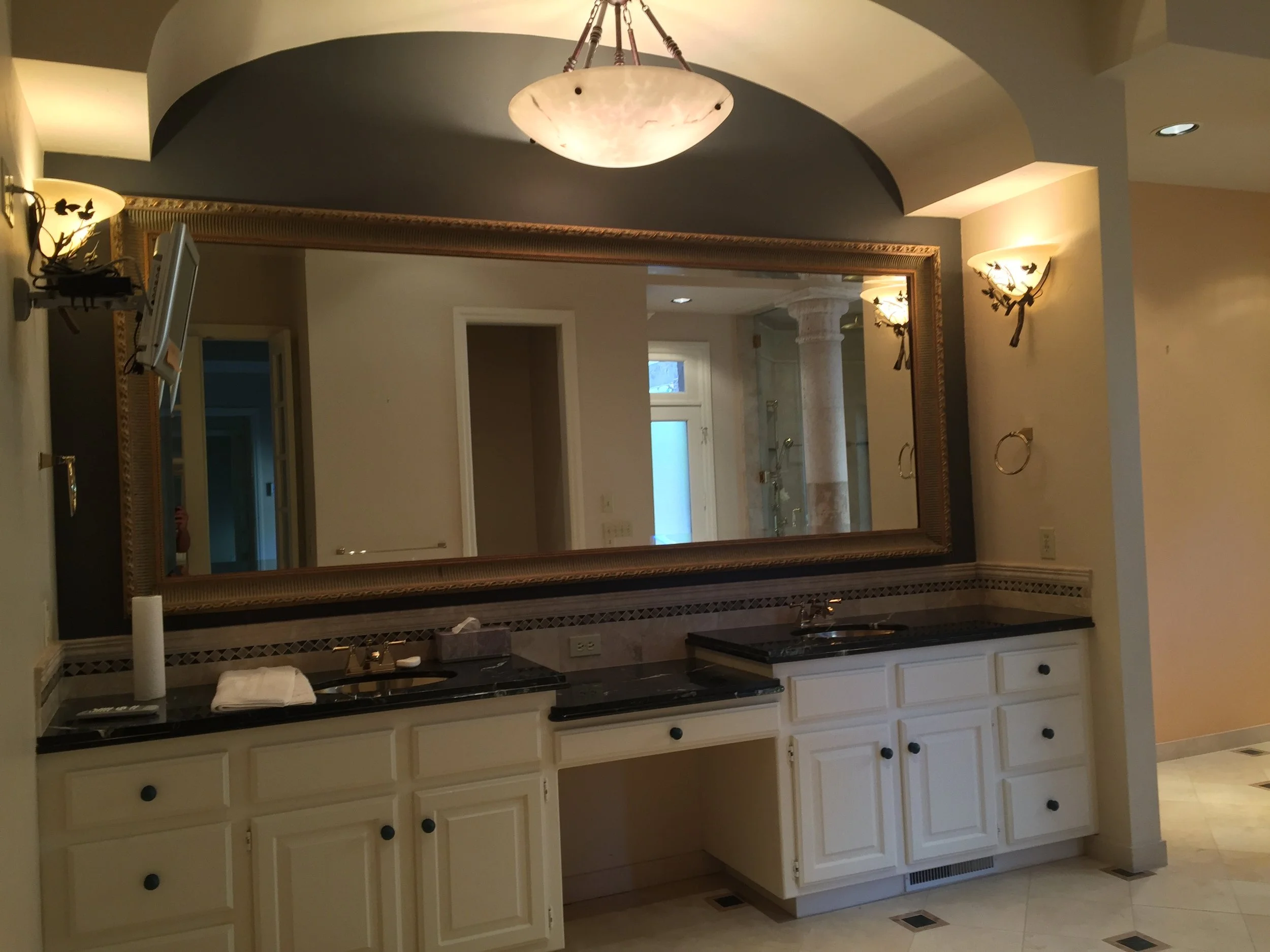 Before renovation: Double vanity with white raised-panel cabinetry, black countertops, large framed mirror, decorative wall sconces, arched ceiling detail, vessel sinks, and recessed makeup station.