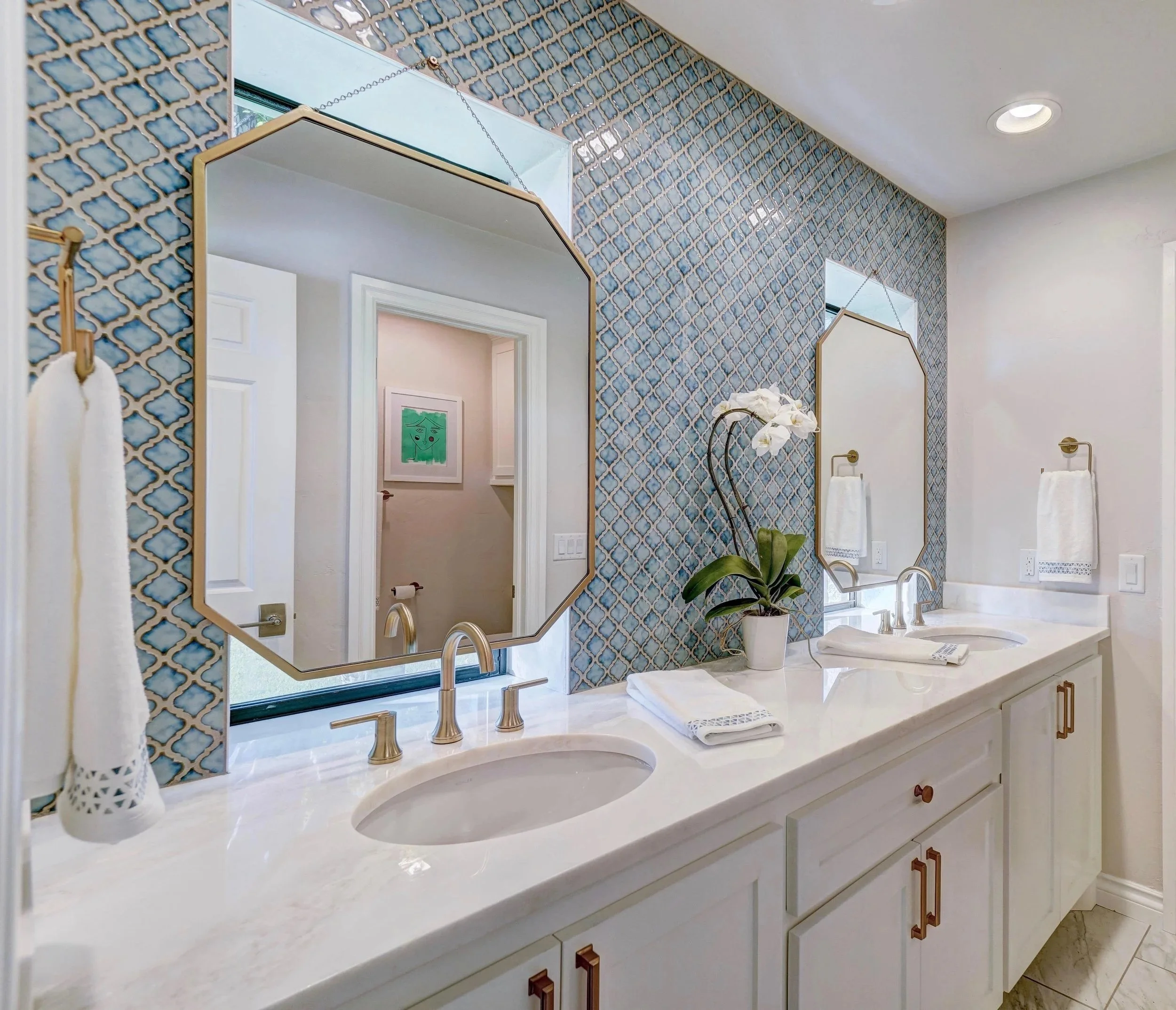 After renovation: Double vanity with white cabinetry, quartz countertop, undermount sinks, brushed nickel faucets, geometric blue tile feature wall, hanging mirrors, brass hardware, and recessed lighting.