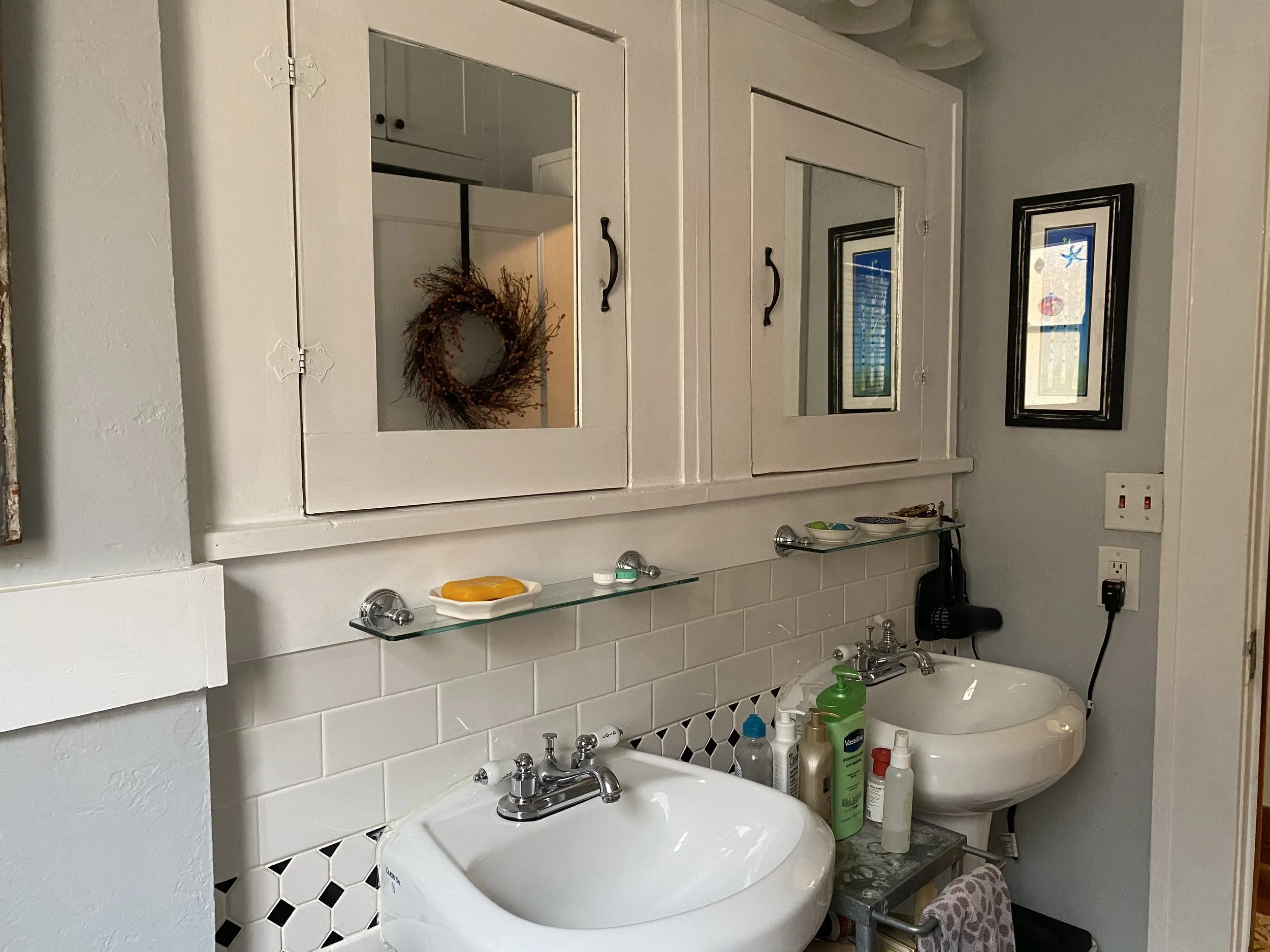 Before renovation: Double wall-mounted sinks with chrome faucets, mirrored medicine cabinets, glass shelves, white subway tile backsplash, patterned accent tile, painted walls, and exposed storage below sinks.