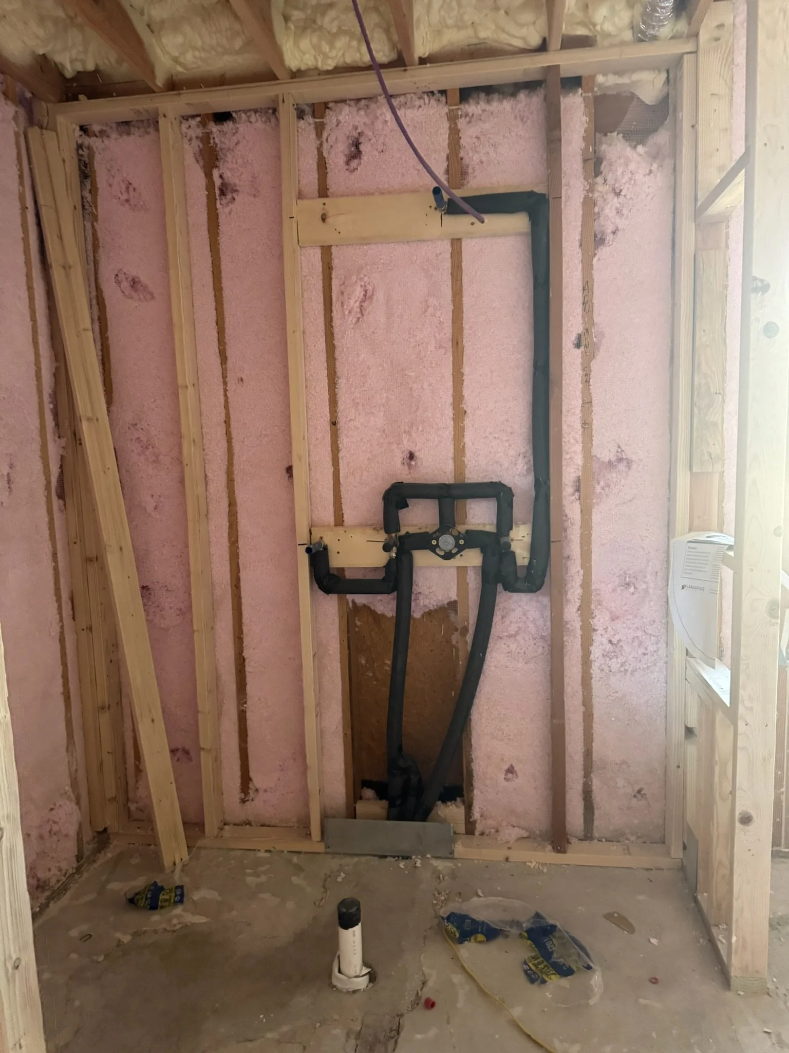 Bathroom demolition showing exposed wall framing, spray foam insulation, in-wall plumbing rough-ins, shower valve assembly, drain line, water supply lines, and unfinished concrete slab prior to construction.